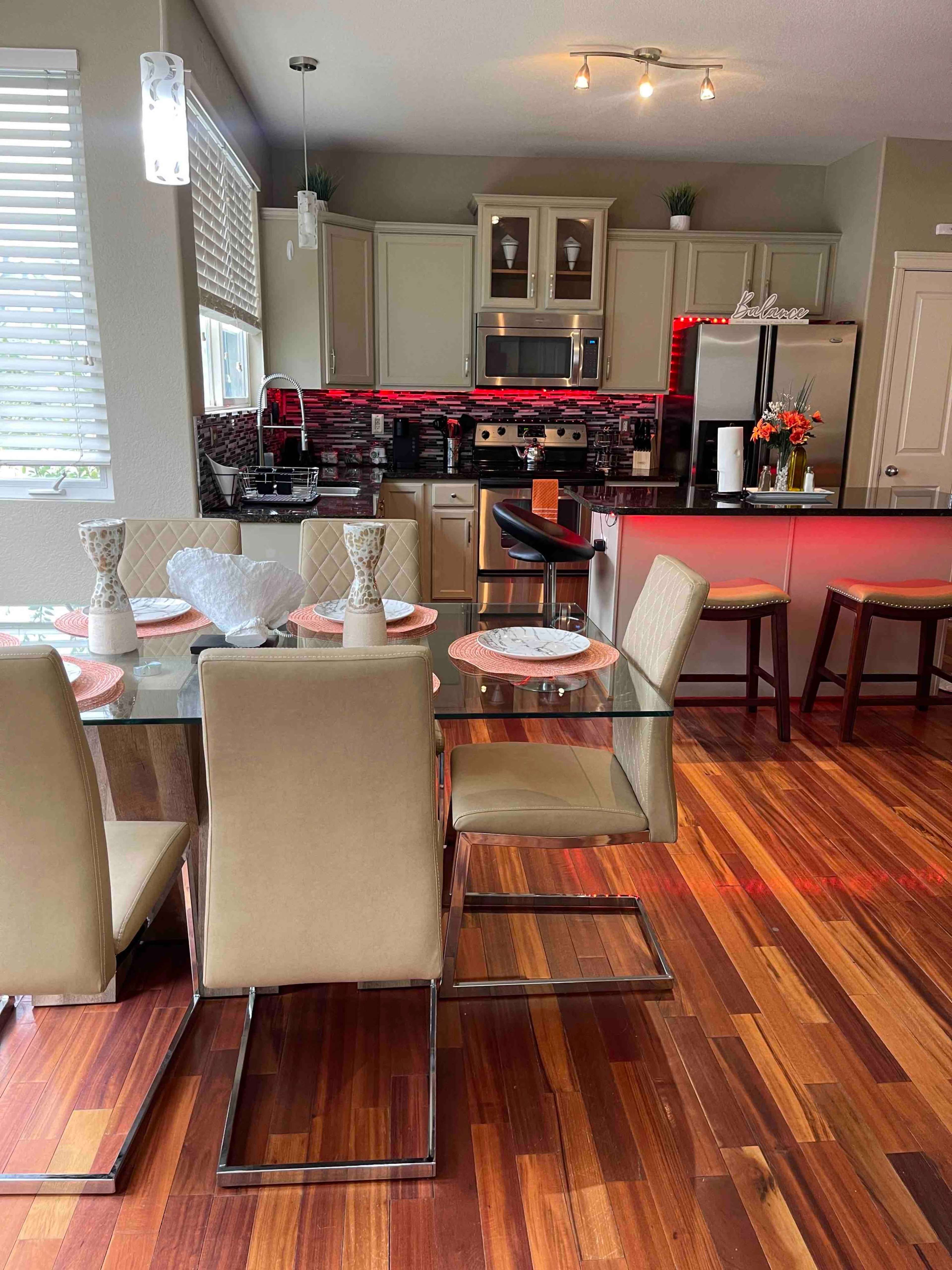 The image shows a modern kitchen and dining area featuring a glass dining table with beige chairs, wooden flooring, and contemporary cabinetry with stainless steel appliances.