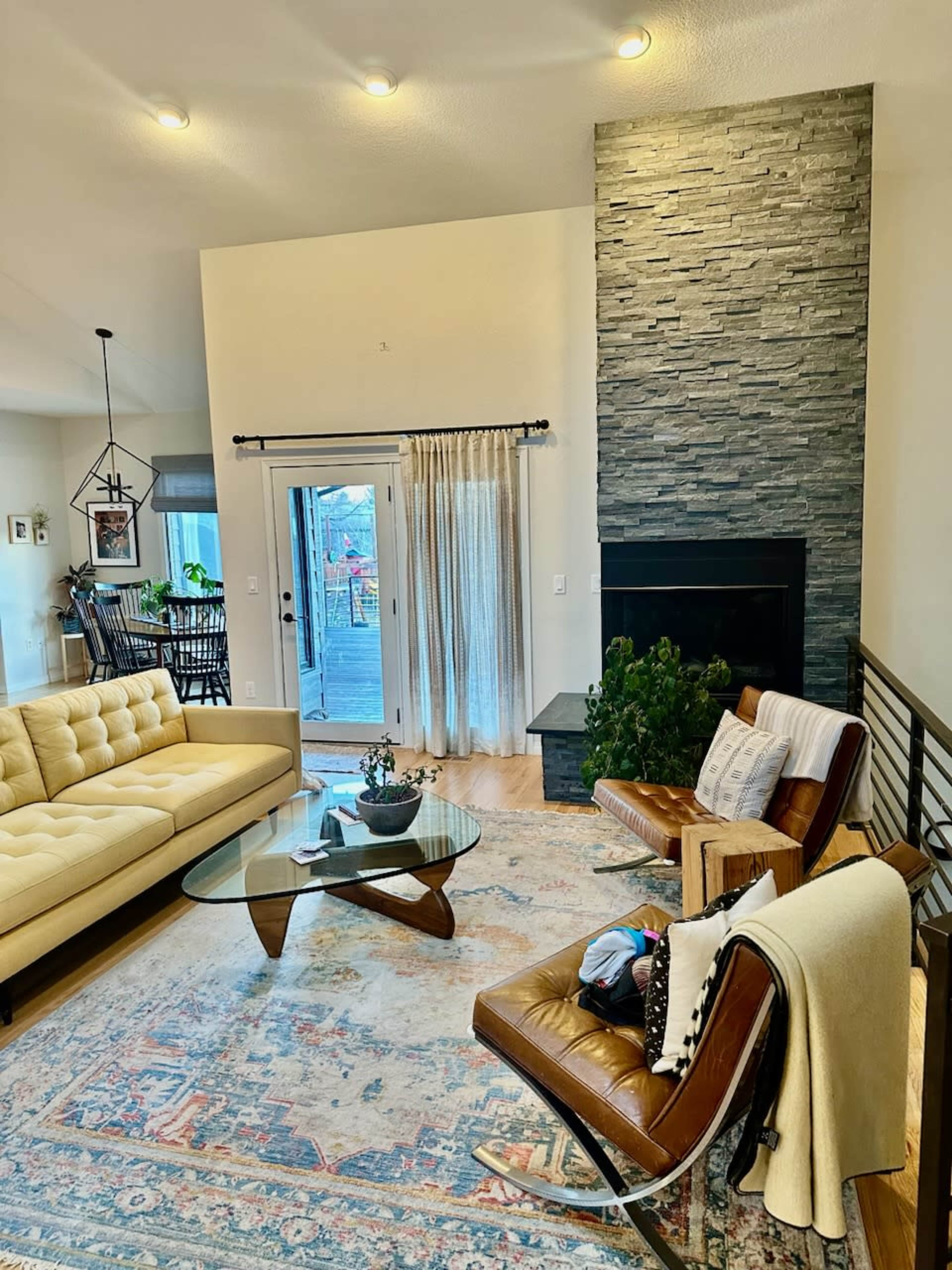 A modern living room with a beige sectional sofa, a glass coffee table, and a stone fireplace, featuring large windows that open to a patio.