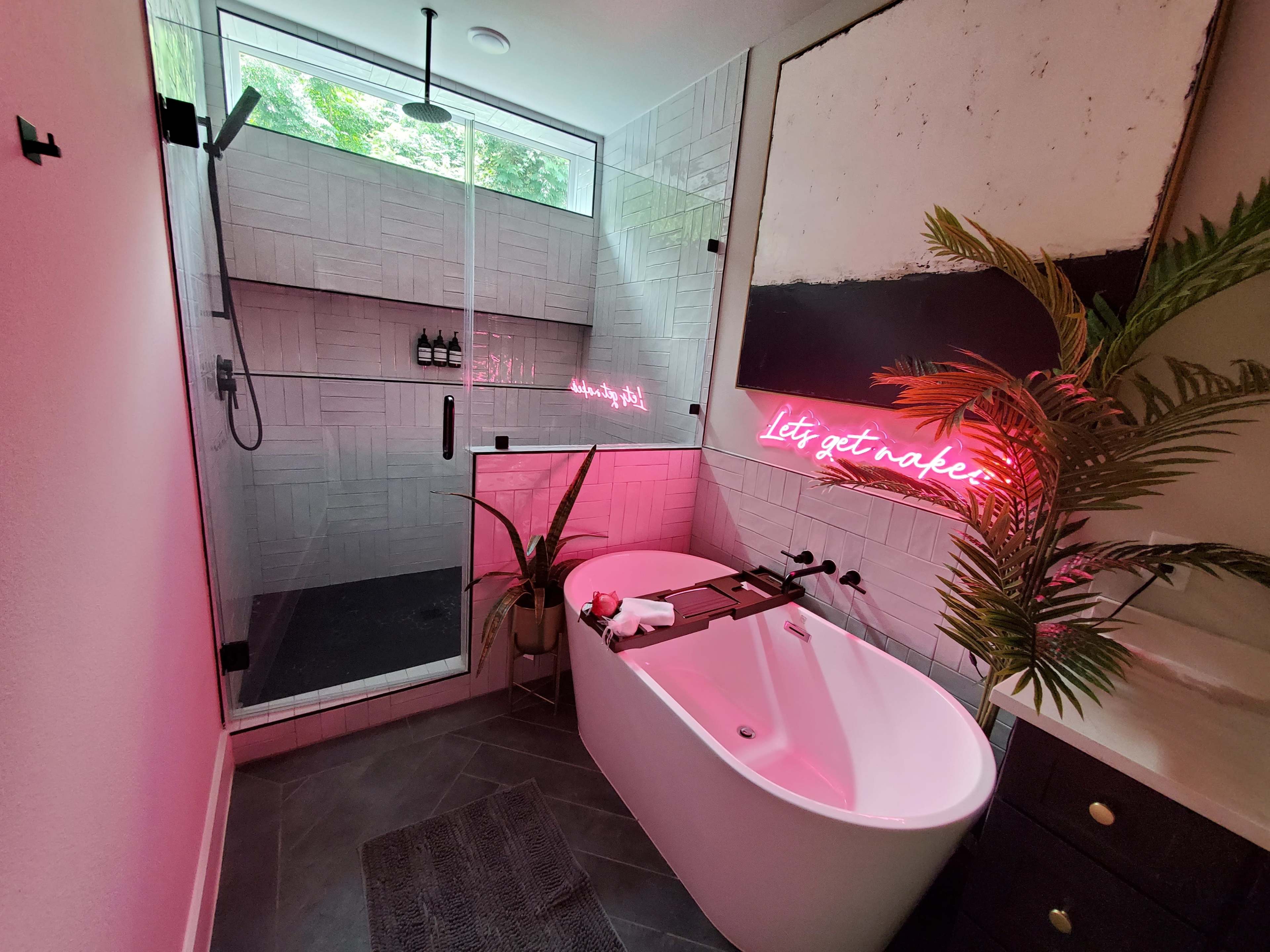 A modern bathroom featuring a freestanding bathtub, a walk-in shower with glass doors, a potted plant, and a neon sign reading "Let's get naked."