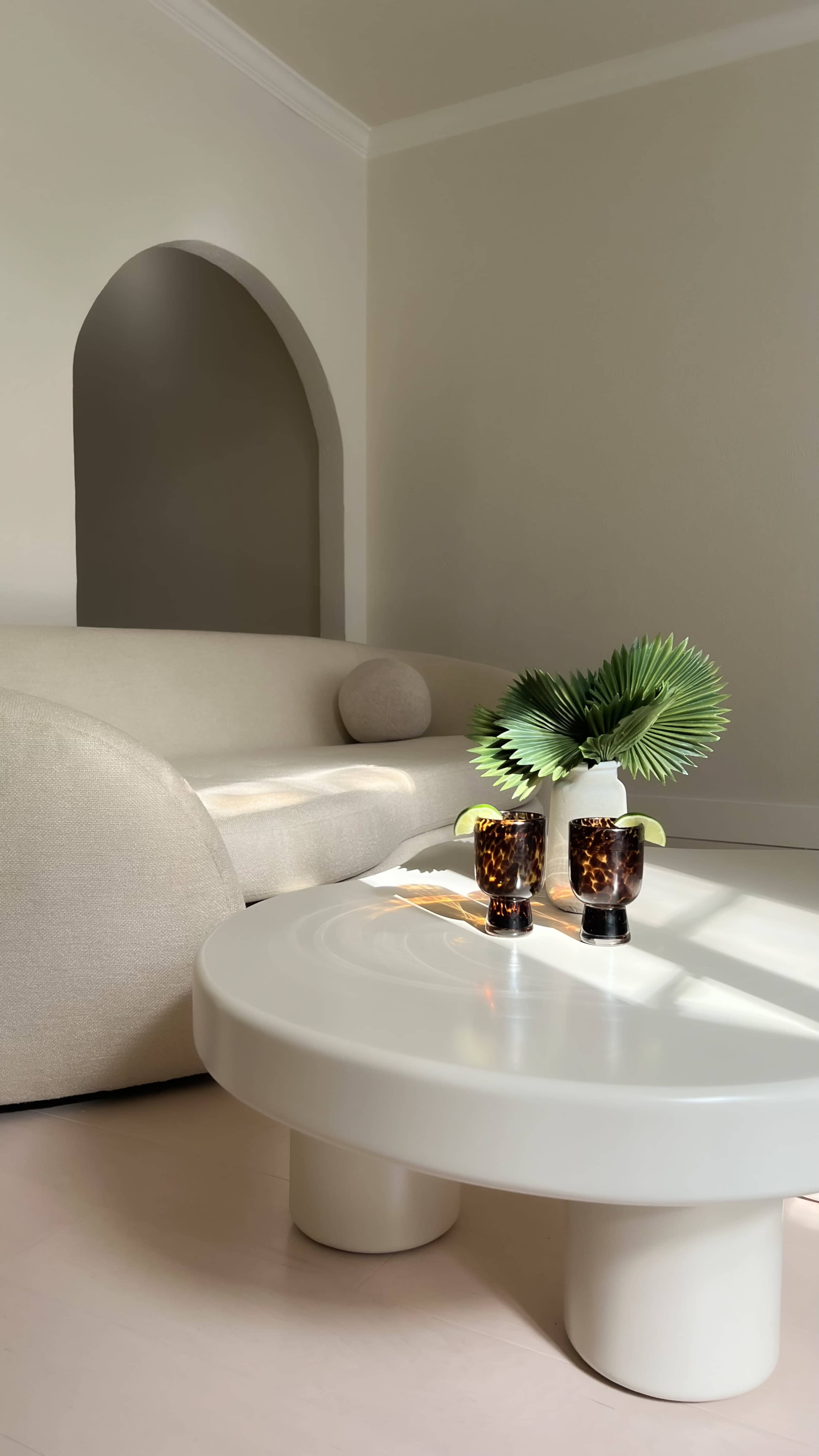 A light-colored living room features a curved sofa, a round coffee table with two patterned glasses, and a potted plant in the center.