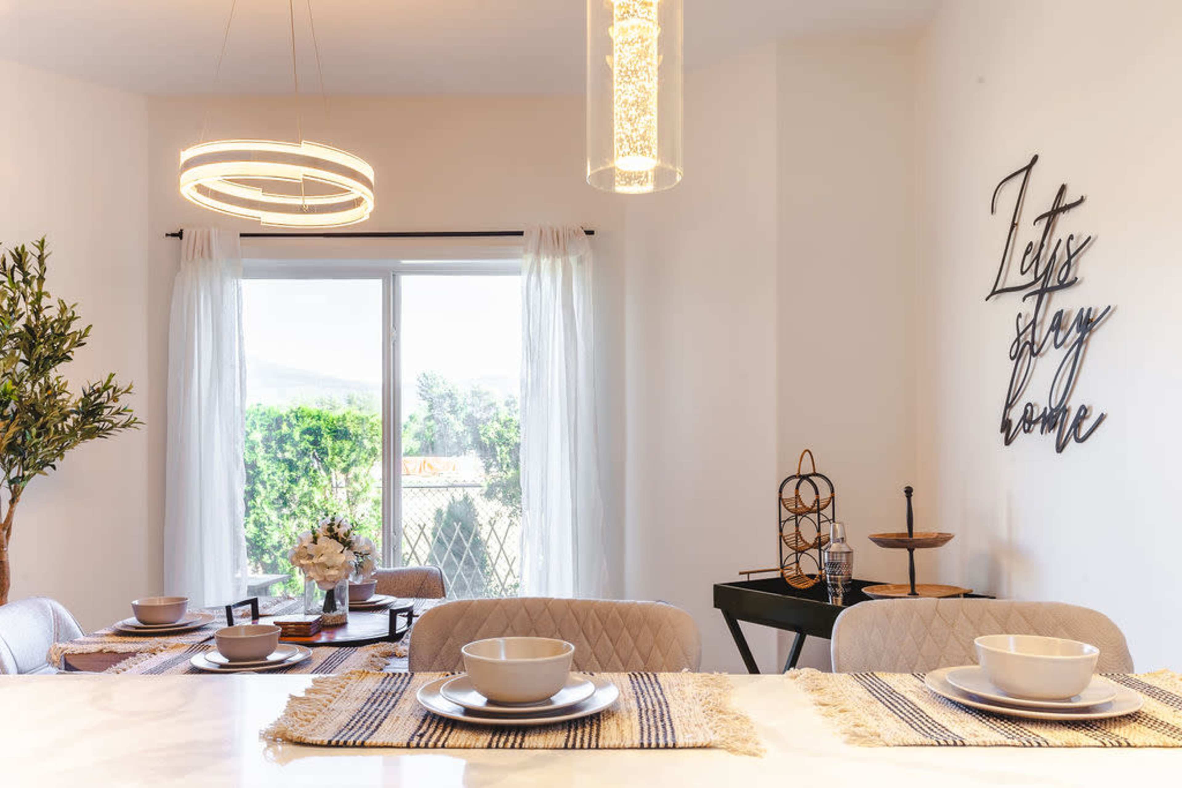 A bright dining area with a modern table set for four, featuring decorative tableware, and large windows overlooking greenery.