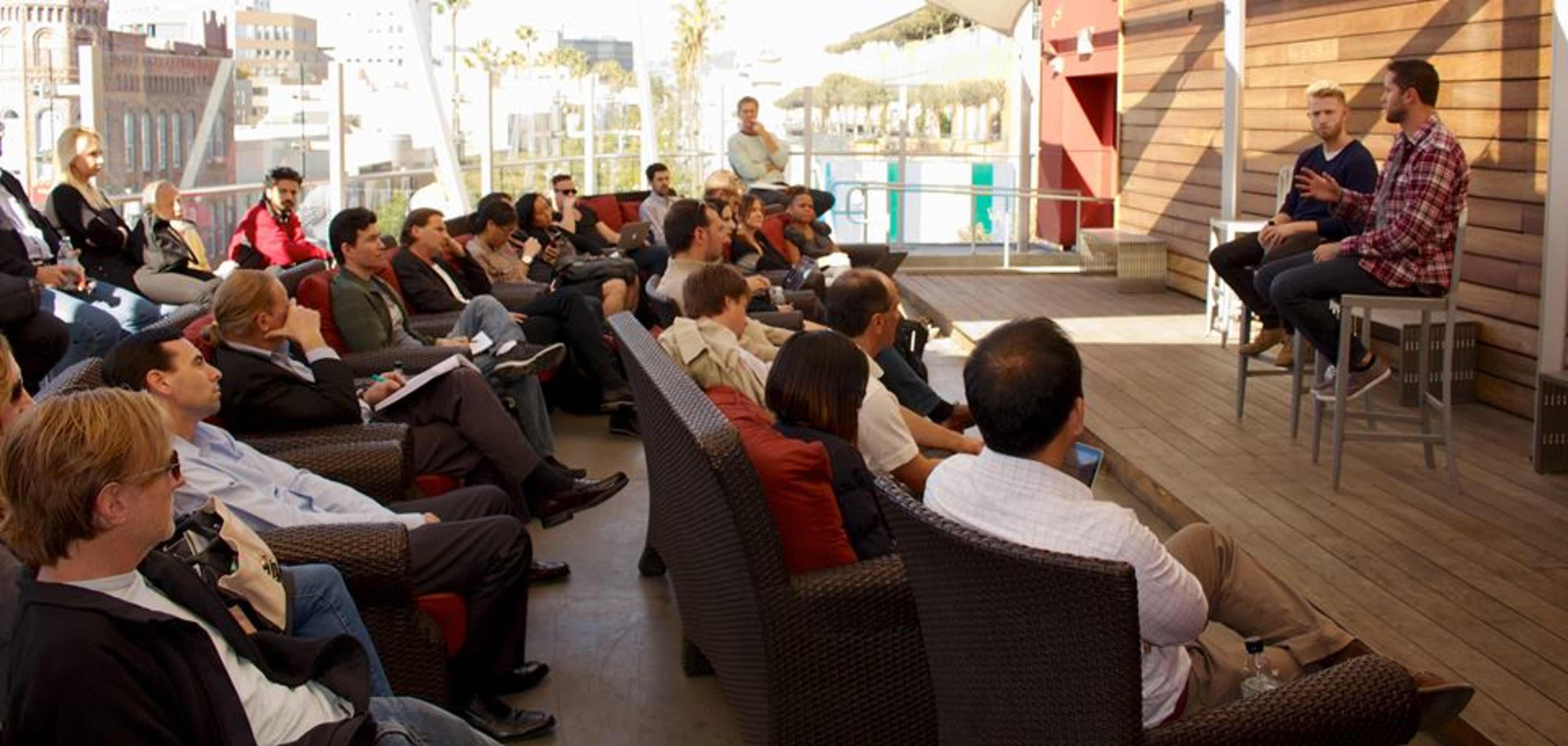 A group of people sits in a tiered outdoor space, attentively watching two speakers engaged in conversation on a stage.