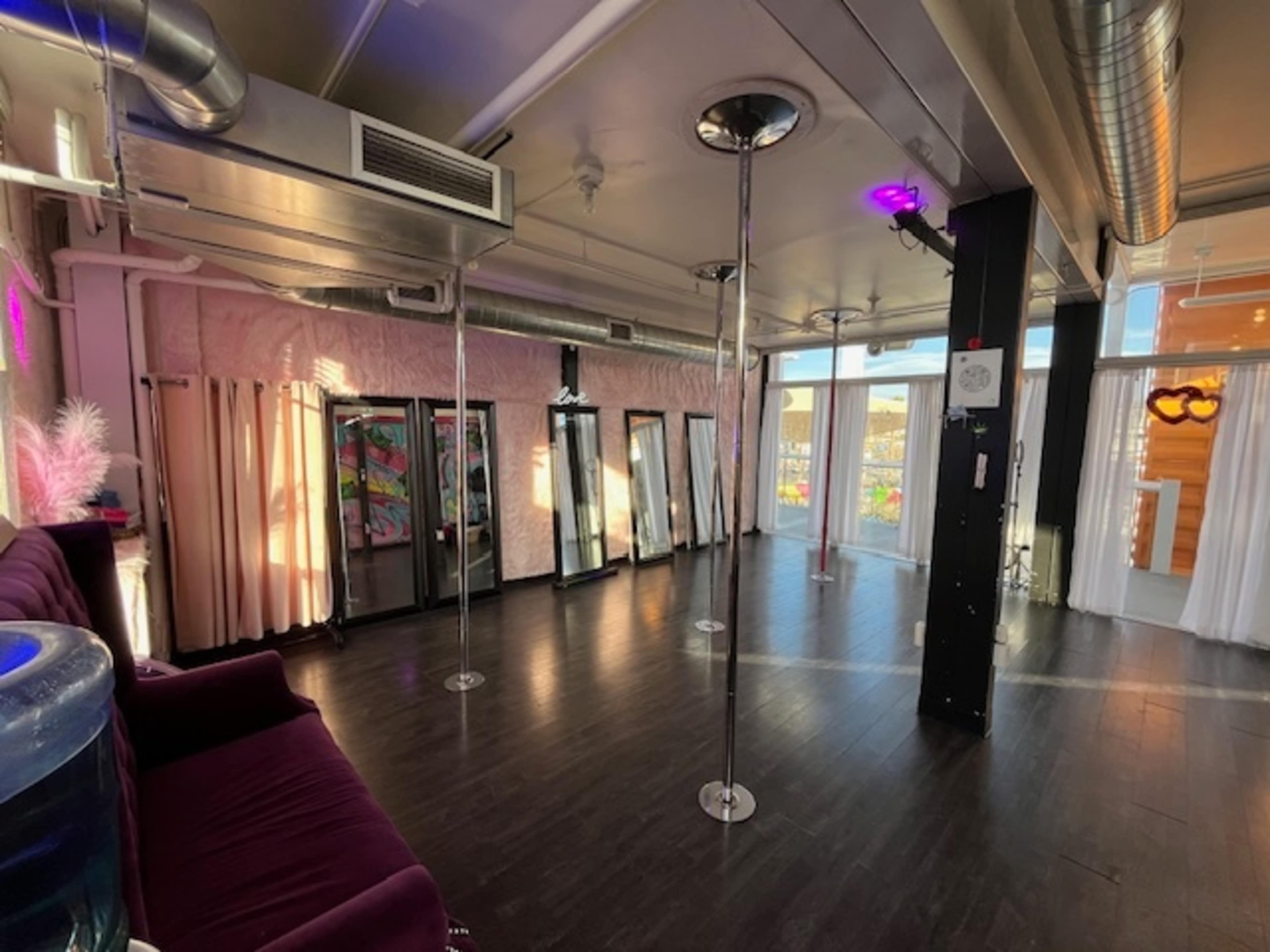 The image shows an interior space designed for fitness or dance, featuring several dance poles, mirrors, and a seating area with a plush couch.