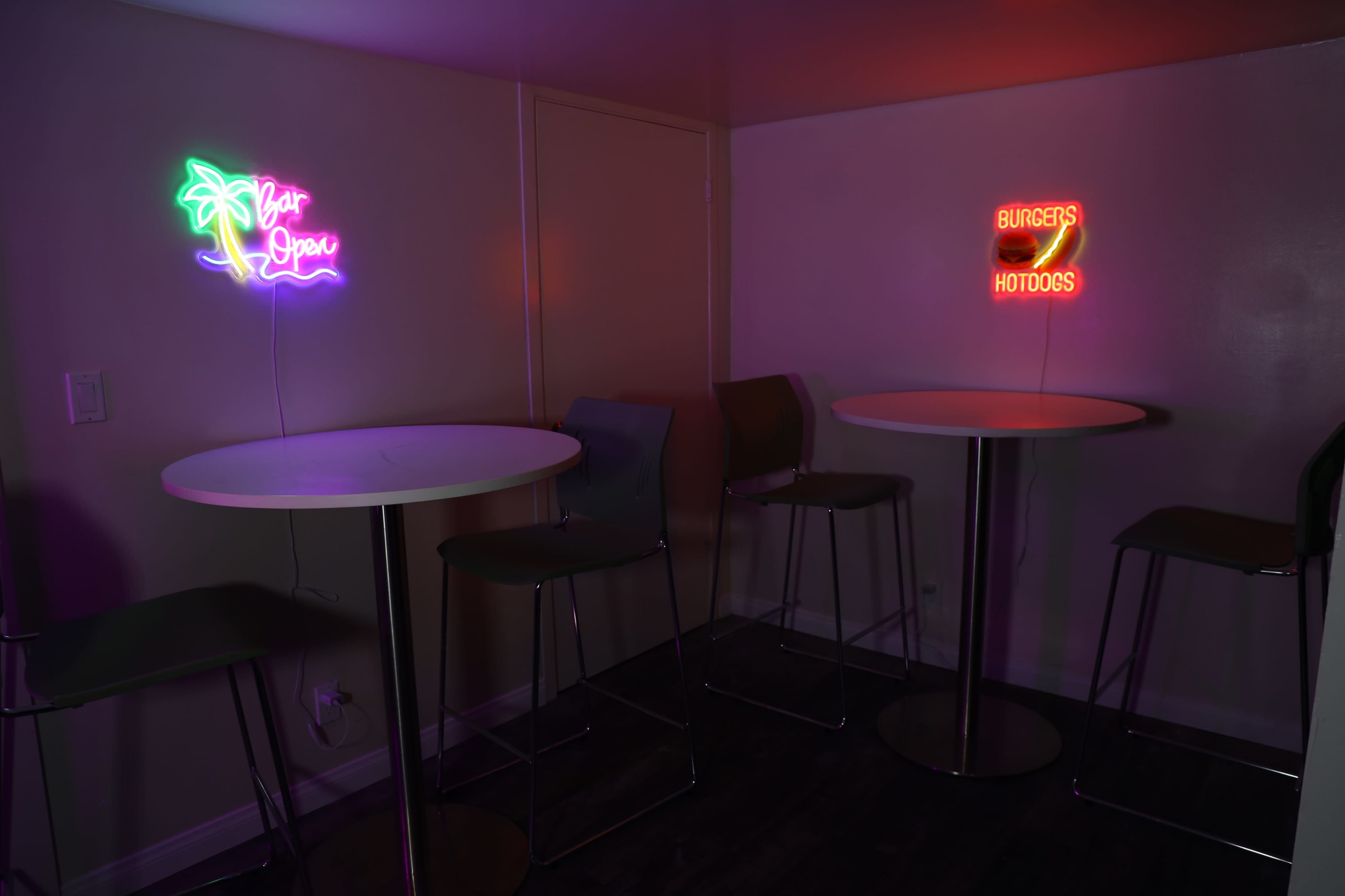 The scene features two illuminated neon signs—one reading "Open" with a palm tree and the other stating "Burgers & Hotdogs"—in a small room with two high tables and chairs.