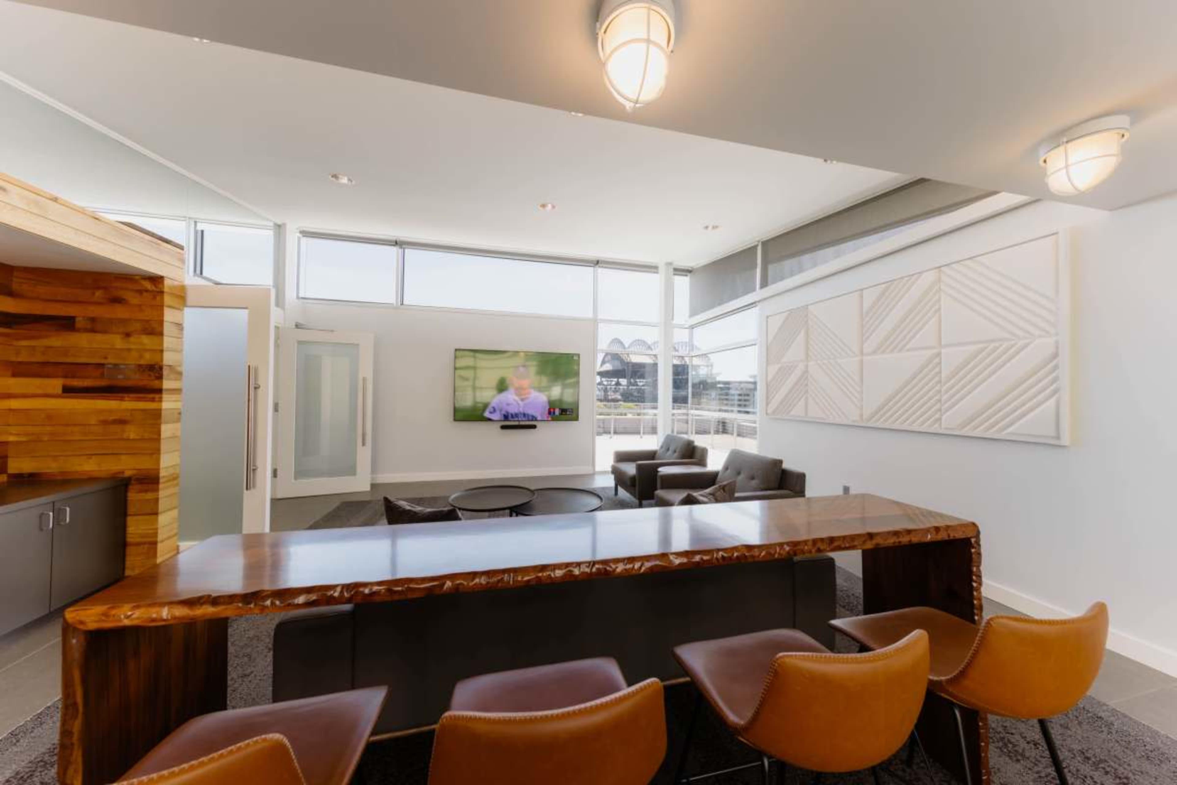 The image shows a modern lounge area featuring a bar with seating, a television mounted on the wall, and large windows letting in natural light.