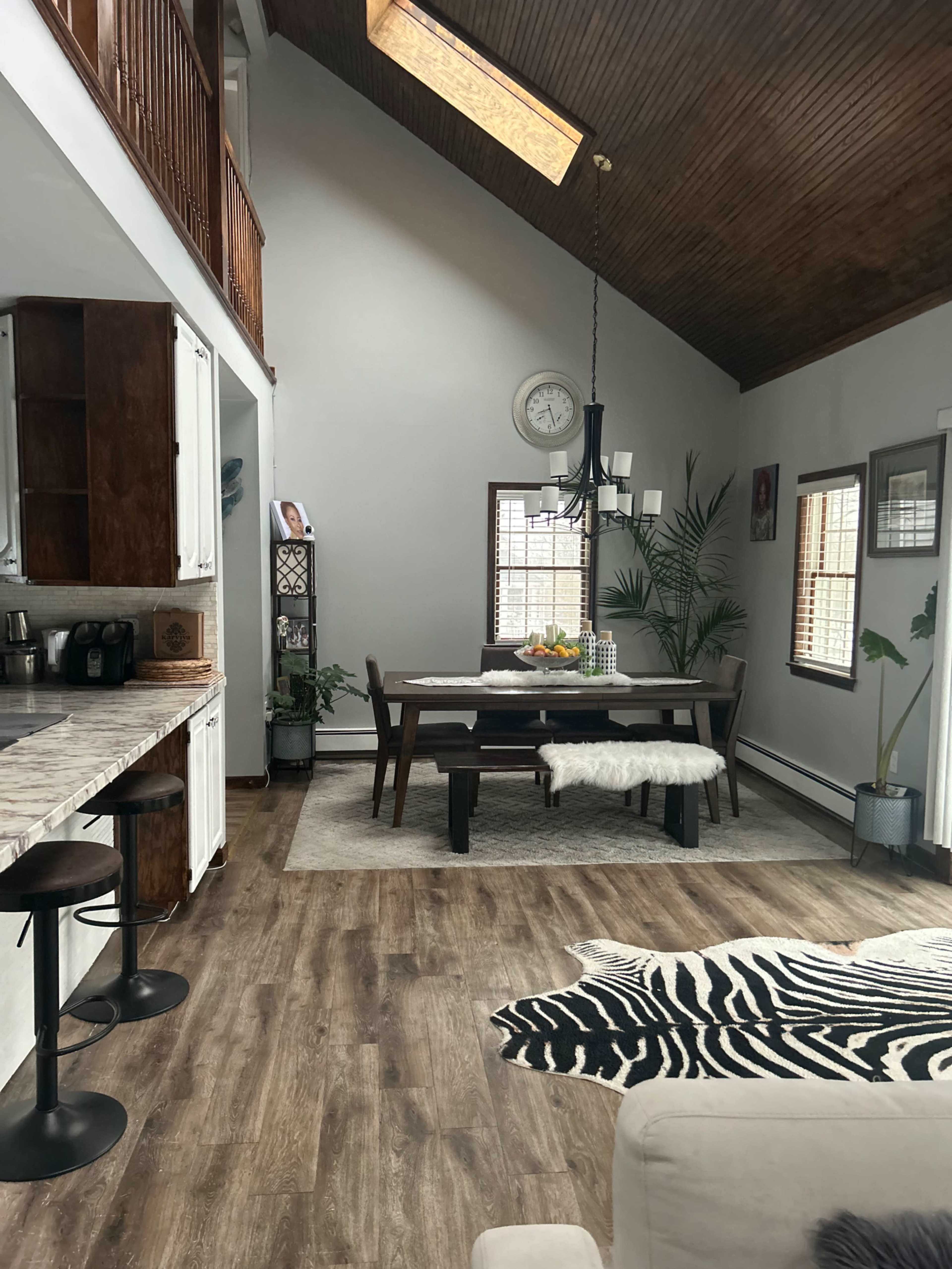 The image shows a modern open-concept kitchen and dining area with high ceilings, featuring a wooden table set for dining and a zebra-patterned rug on the floor.