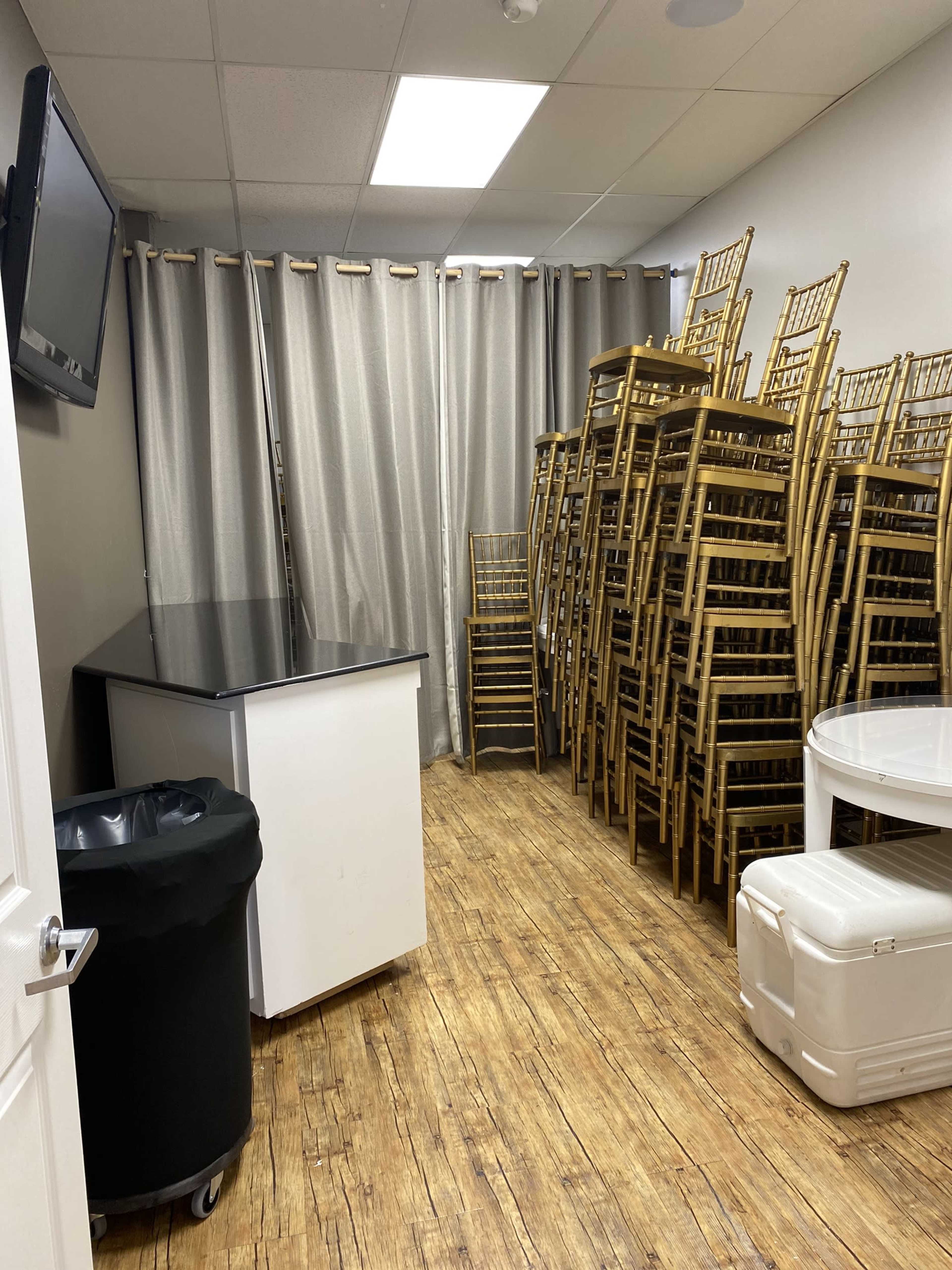 The image shows a small room with a stack of gold-colored chairs piled against a wall, a black trash can, and a few tables in the space.