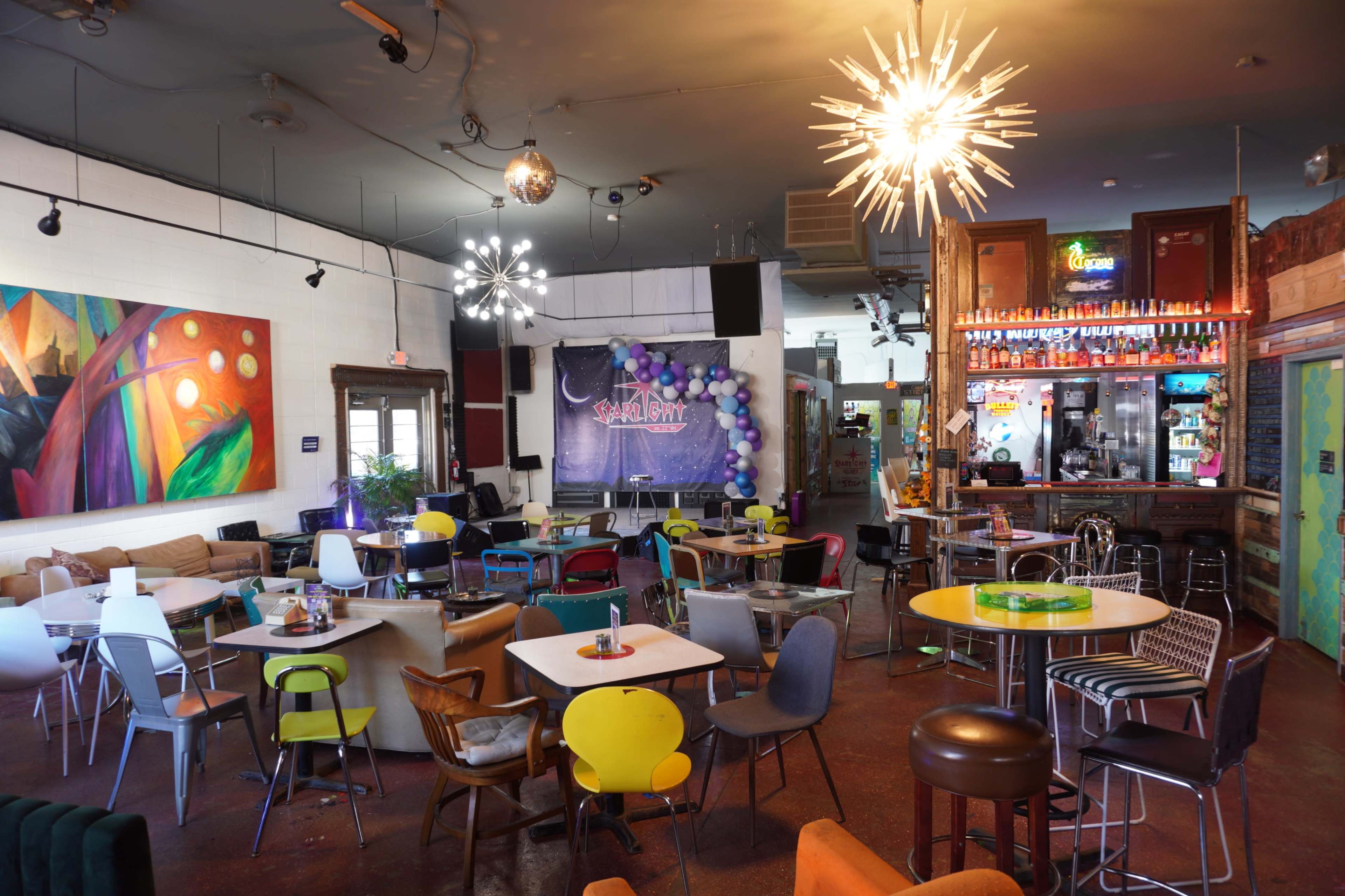 The image shows a lively café with various seating arrangements, colorful chairs, and decorative lighting fixtures.