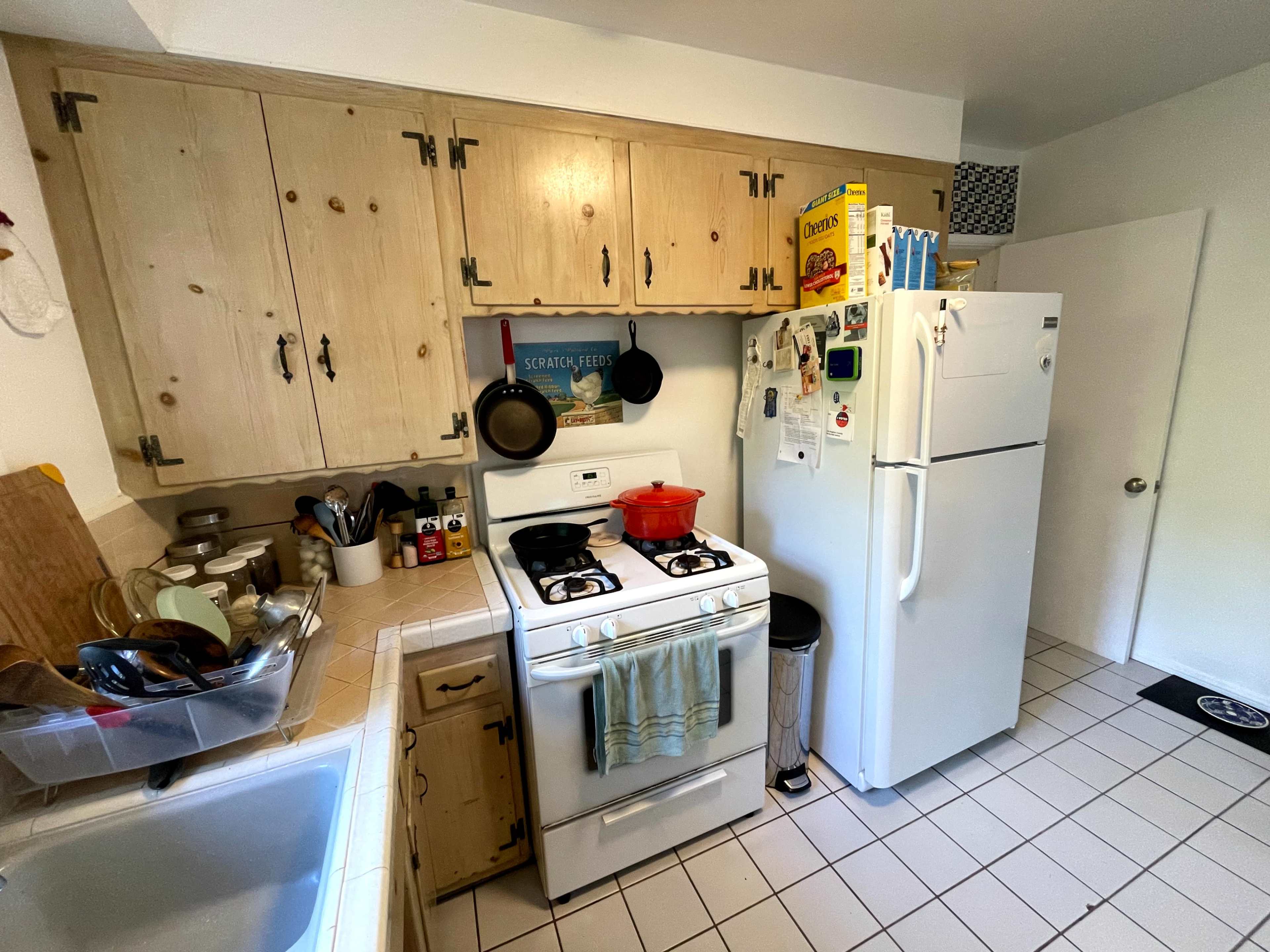 The image shows a small kitchen with wooden cabinets, a gas stove, a refrigerator, and various kitchen utensils arranged on the countertops.