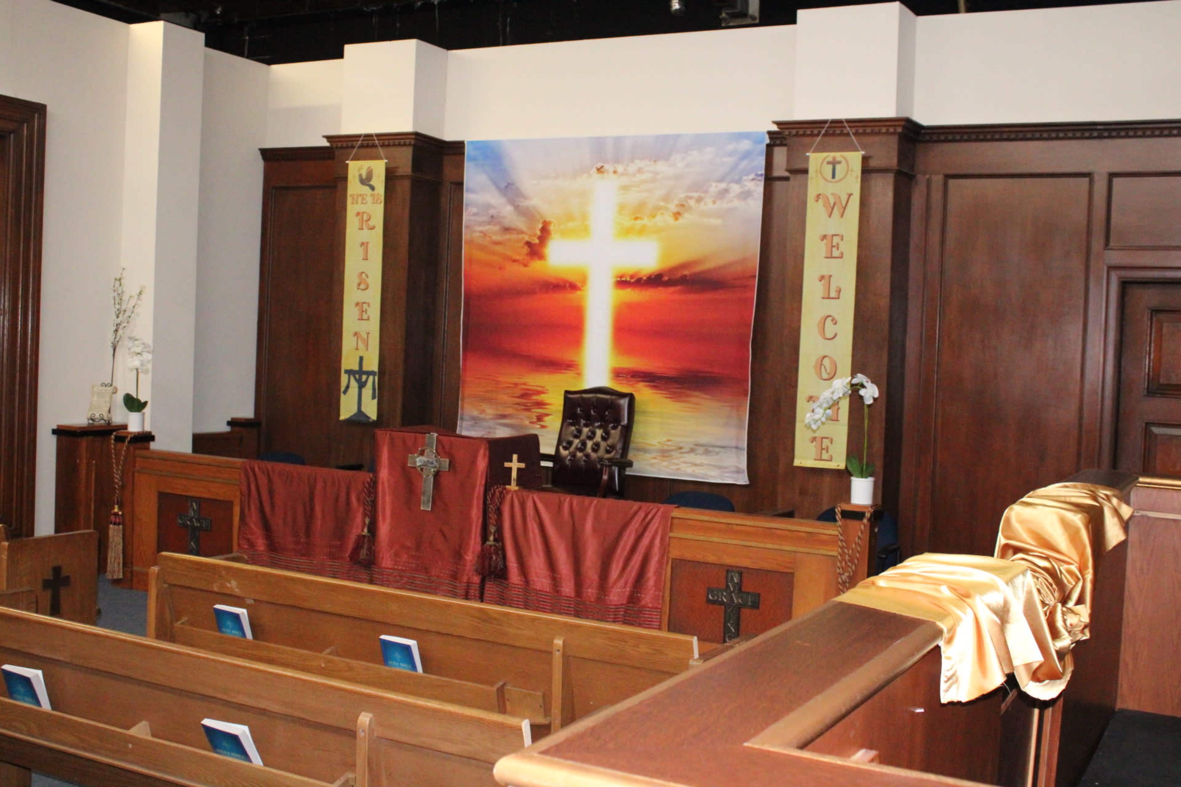 The interior of a church features a central altar with a large backdrop of a cross and sunrise, flanked by banners reading "WELCOME" and "RISEN."