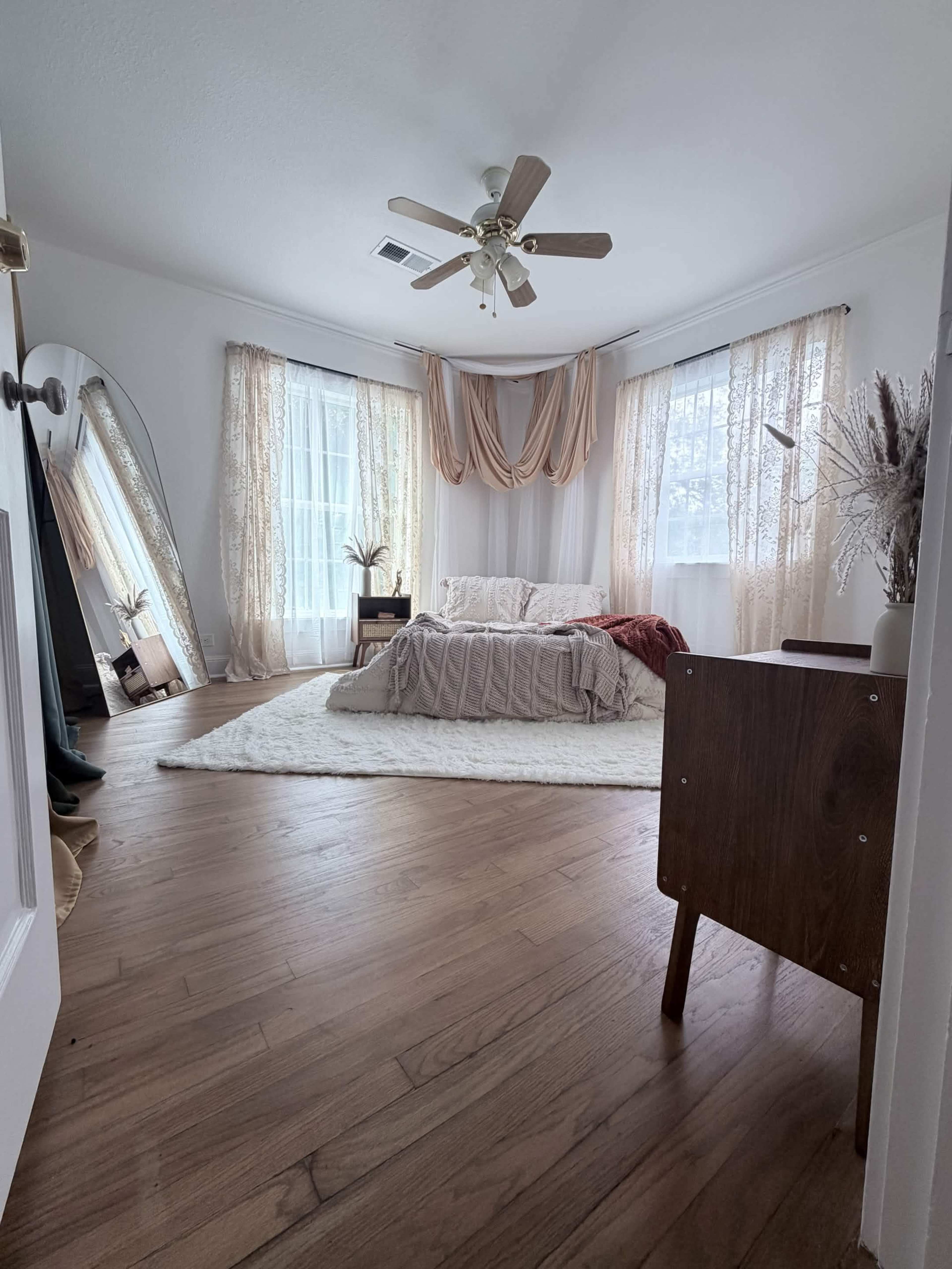 The room features a bed with light bedding, a large mirror, sheer curtains, and wooden flooring, creating a spacious and well-lit atmosphere.