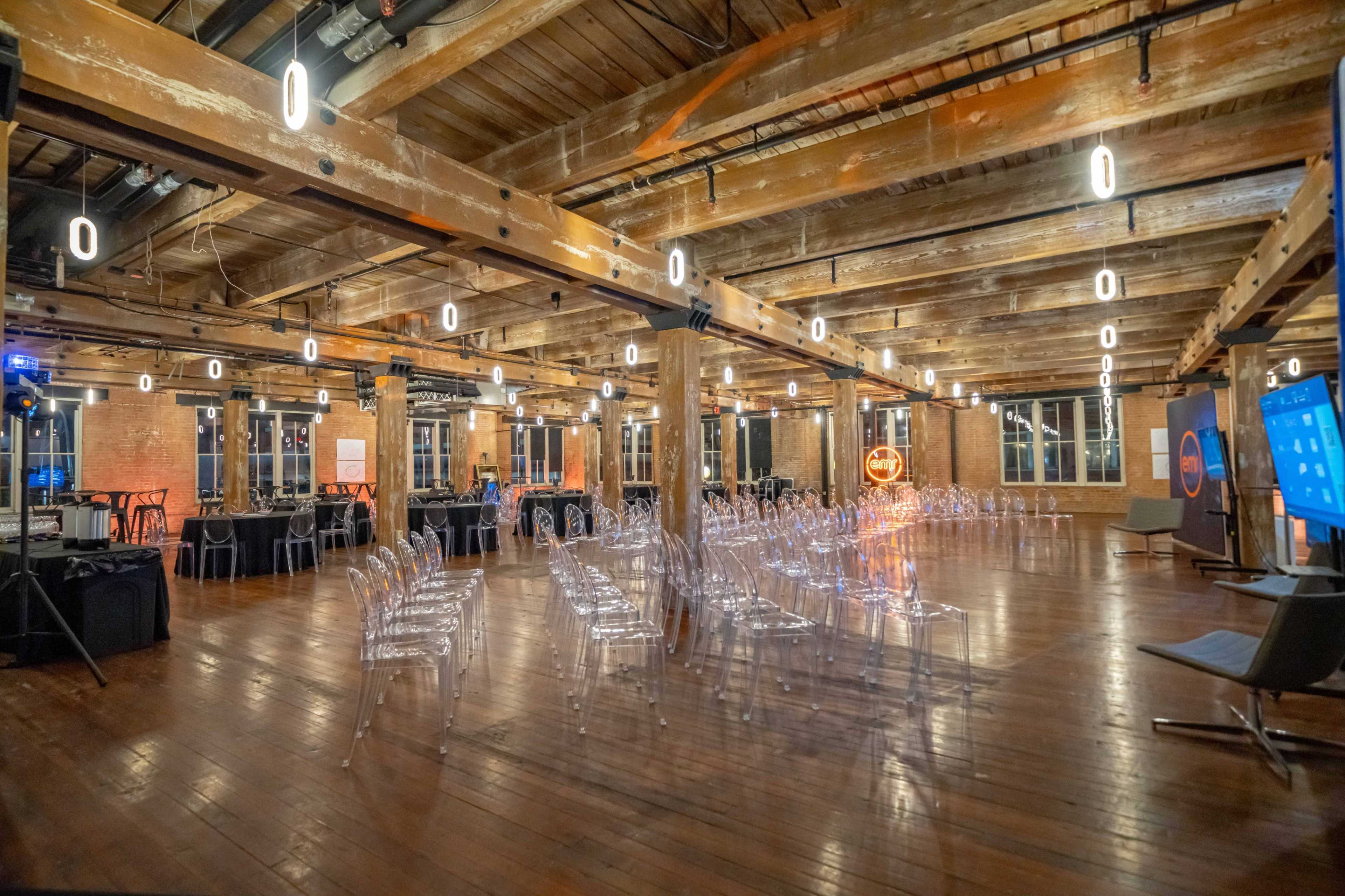The image shows a spacious event venue with wooden beams and floors, set up with clear chairs arranged in rows facing a stage area.