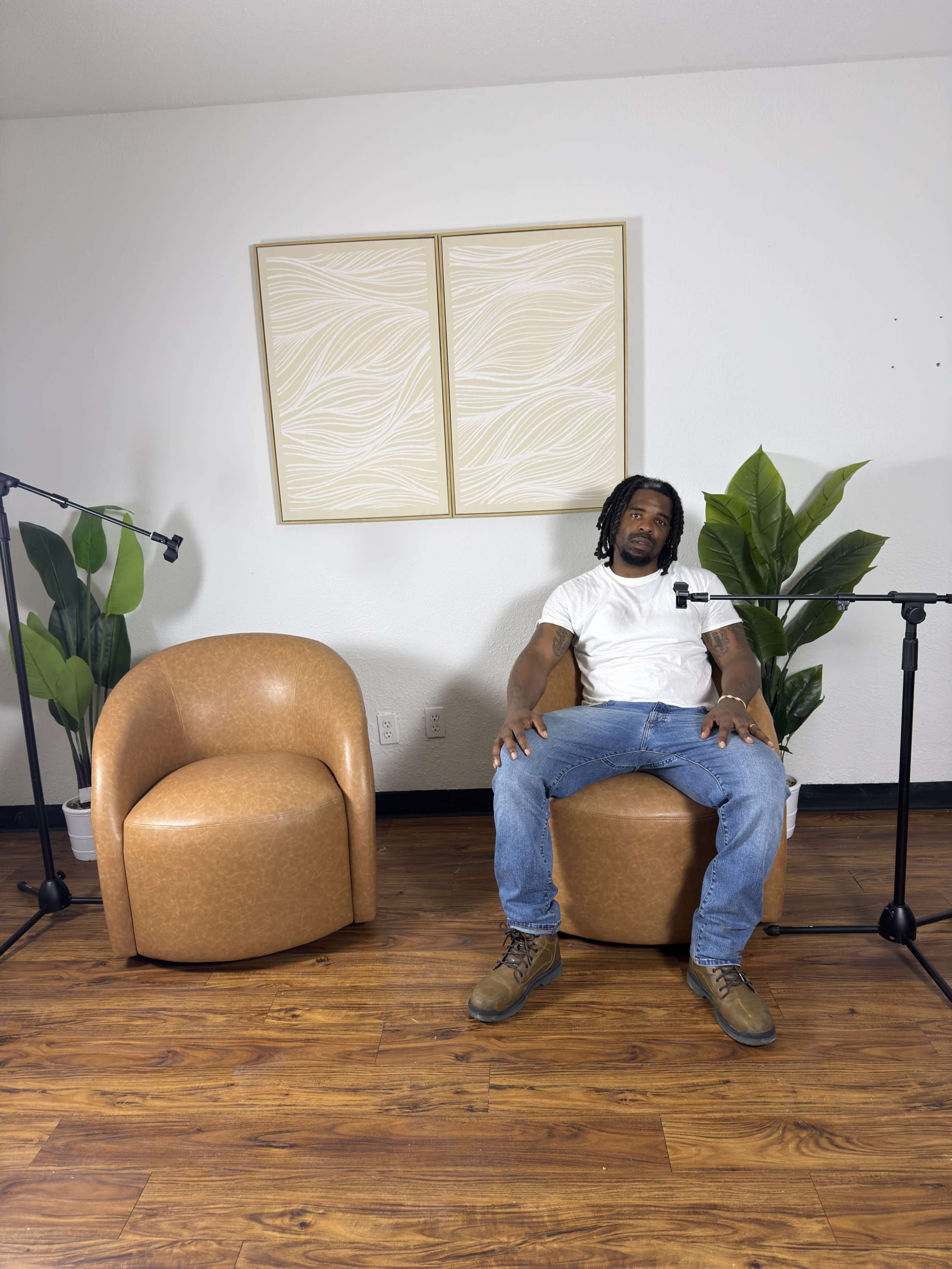 A man is seated on a round chair in a minimalist room equipped with two microphones and a potted plant.