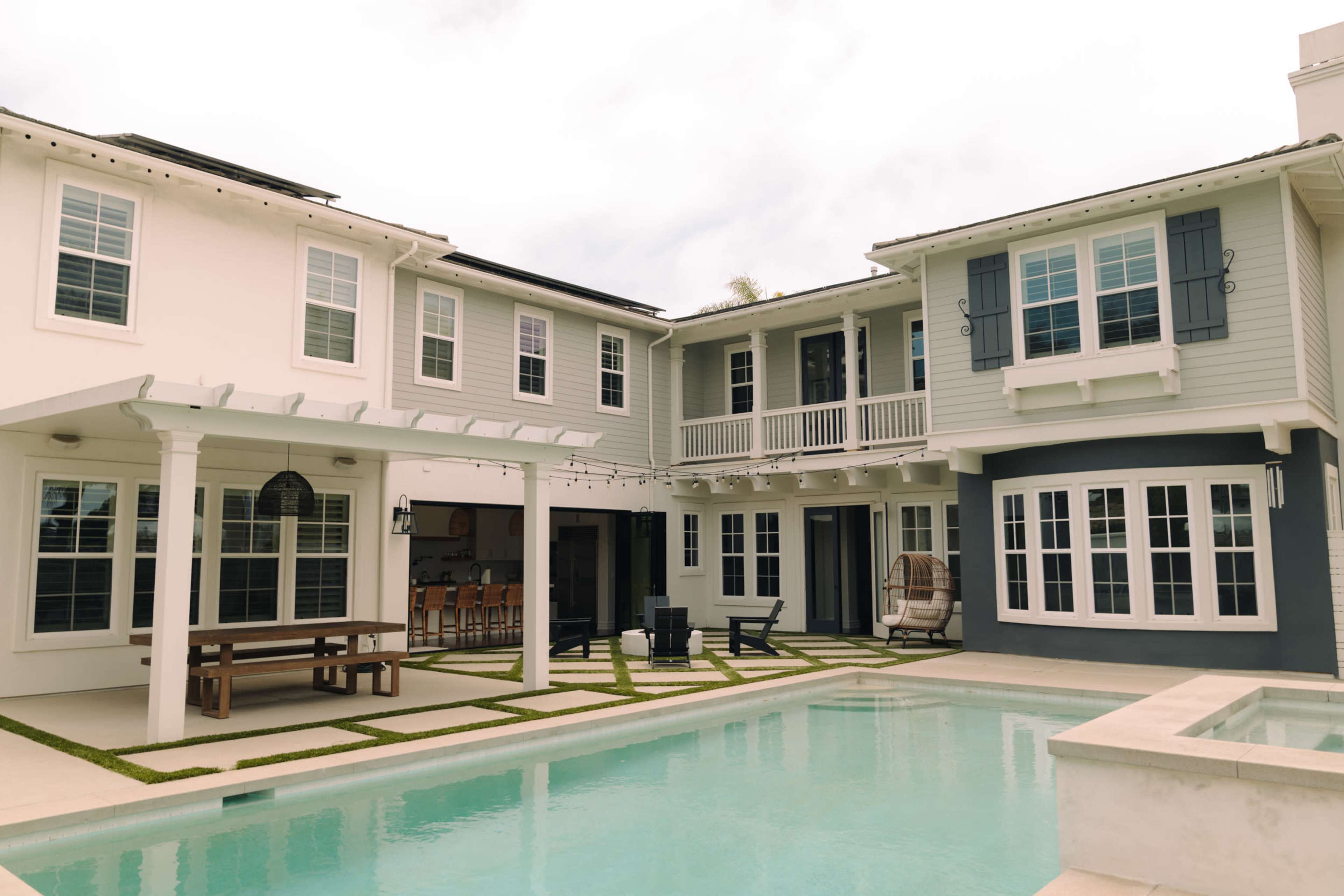 The image shows a modern two-story house with a pool in the backyard, featuring a patio area with outdoor seating and a dining table.