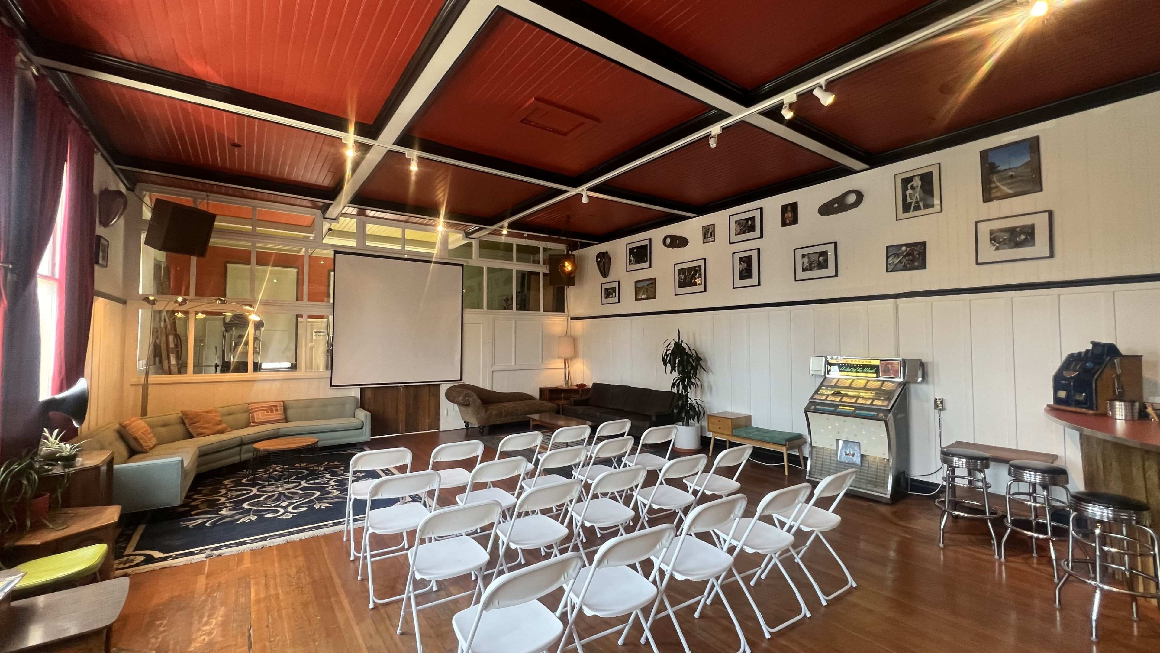 A spacious room set up with rows of white chairs facing a large screen, surrounded by framed photographs on the walls and vintage decor including a jukebox.