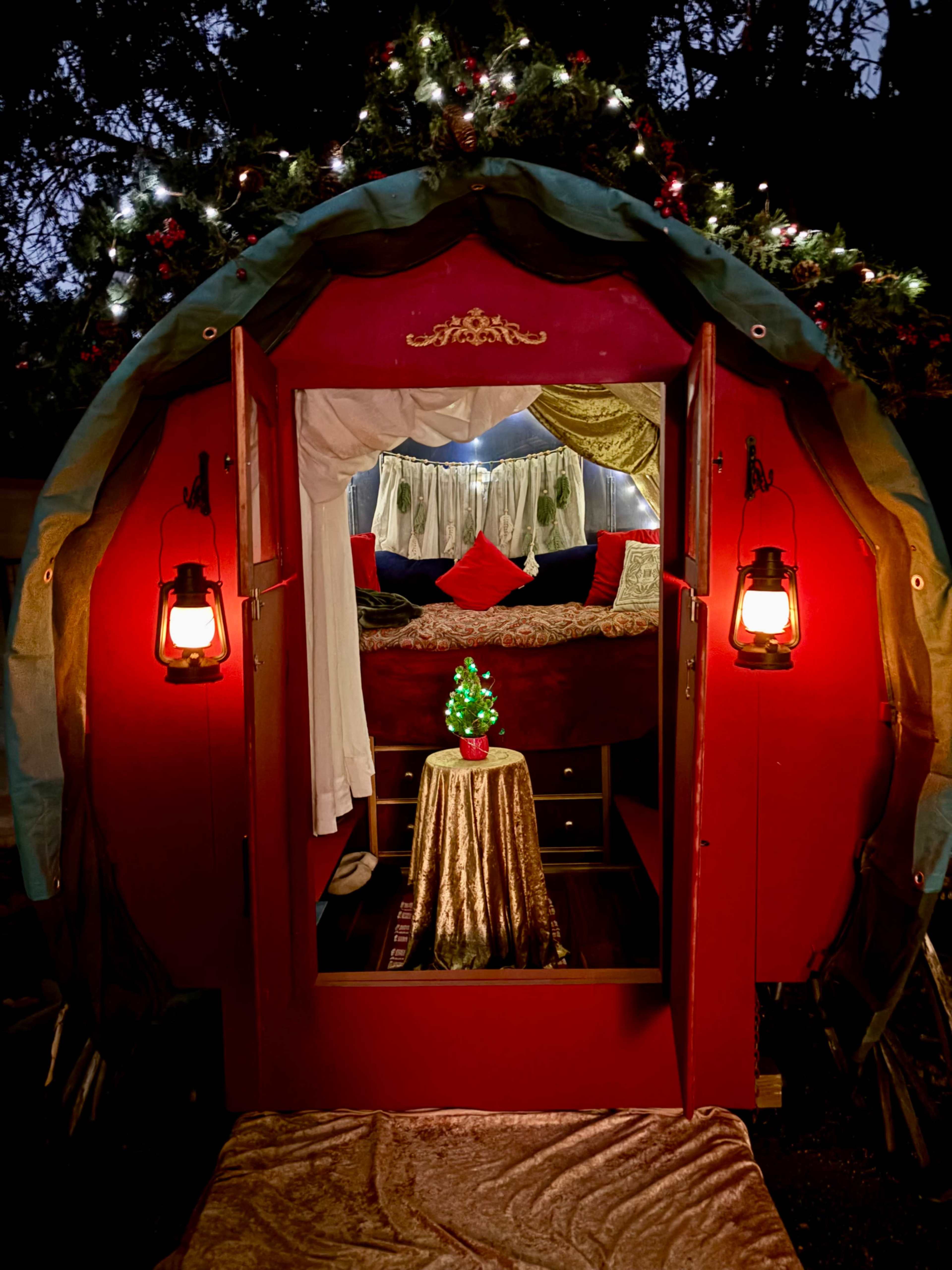 A cozy, red tent with lanterns at the entrance features a decorated interior with a small Christmas tree on a table.