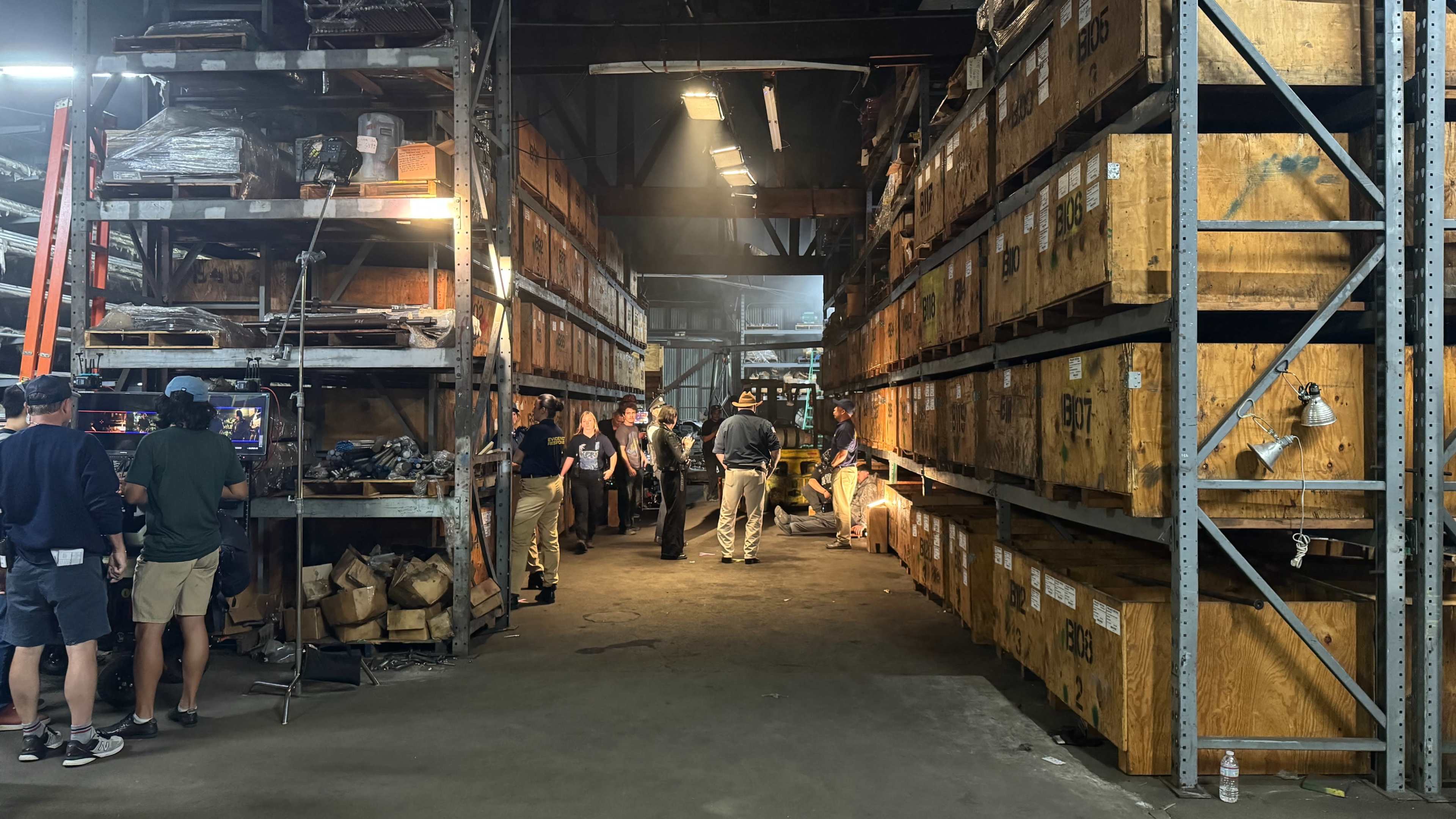 The image shows a warehouse aisle filled with wooden storage crates on either side, with people walking and working amid the stacks.