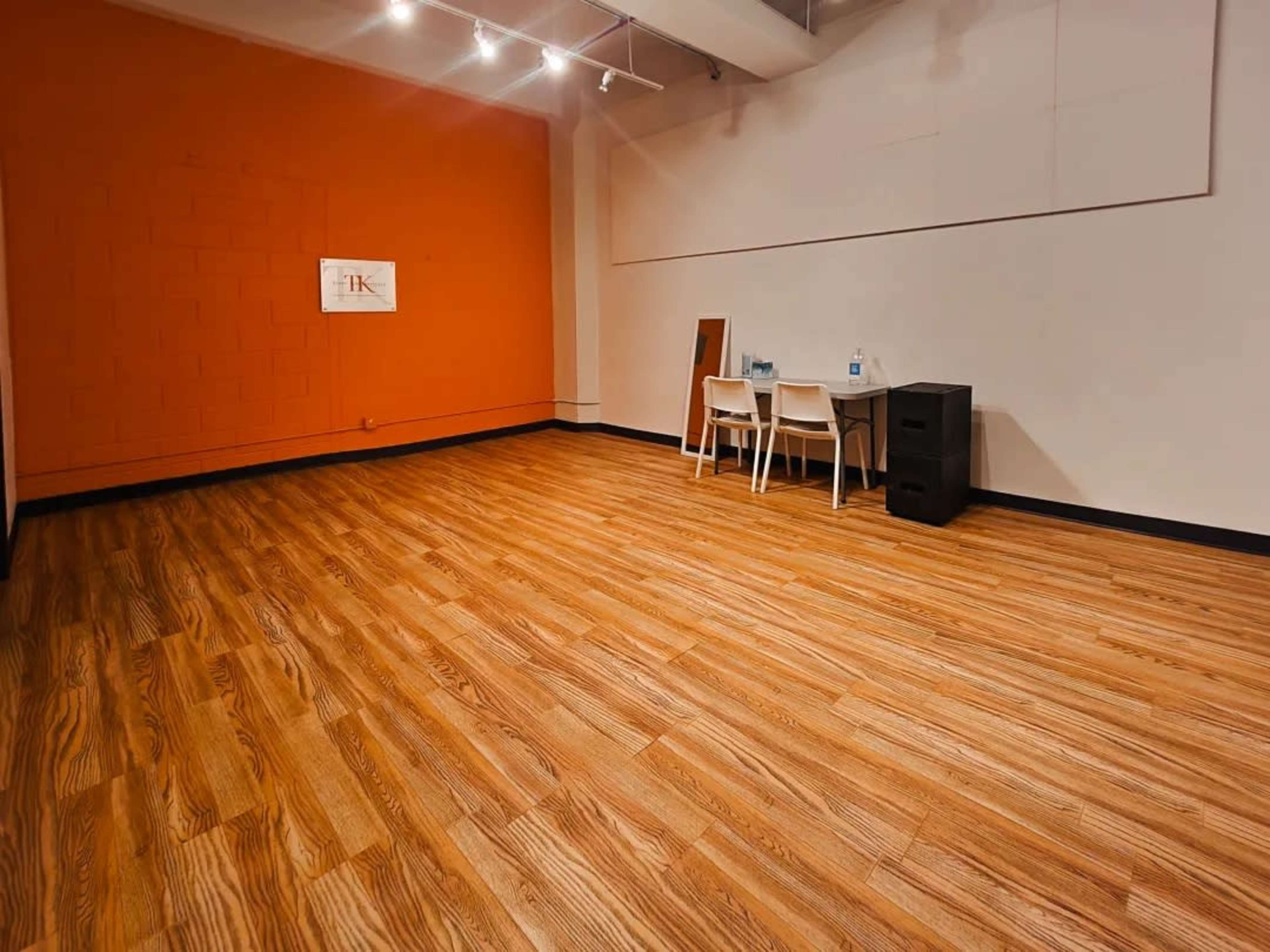 The image shows a spacious room with orange and white walls, wooden flooring, and minimal furniture, including a table with three chairs and a small black cabinet.