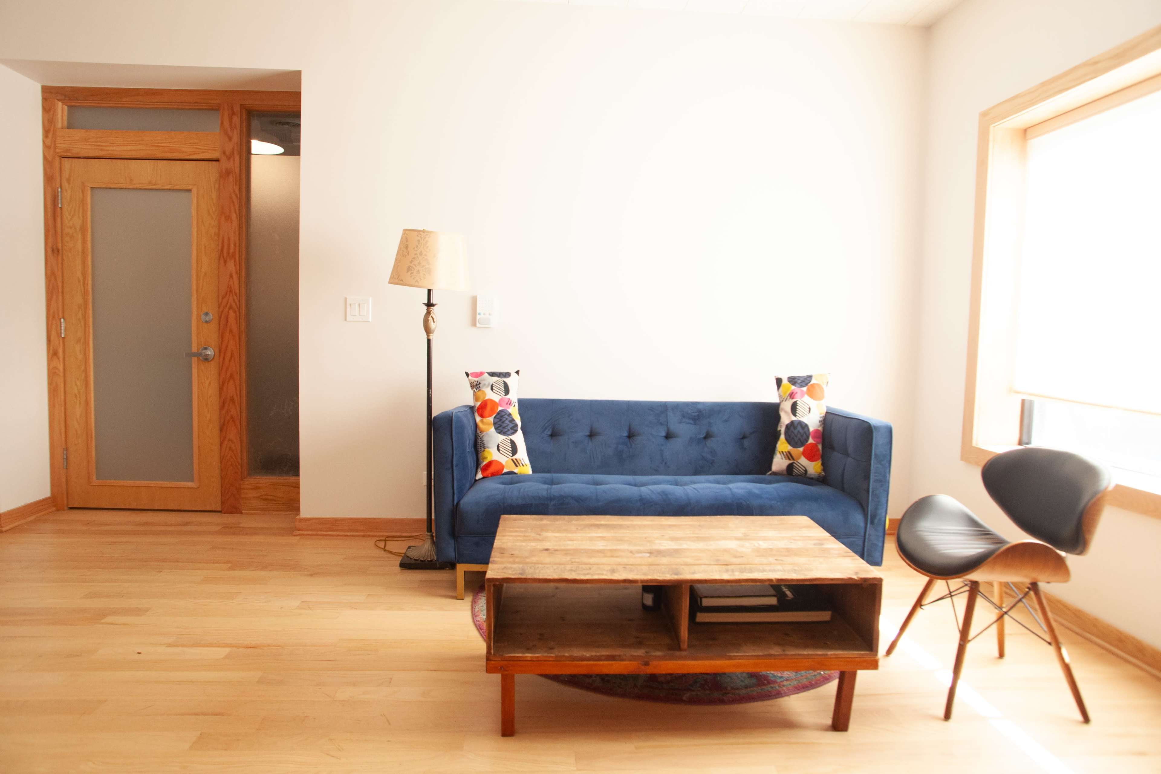 A living room featuring a blue tufted sofa with patterned pillows, a wooden coffee table, and a chair in front of a large window.