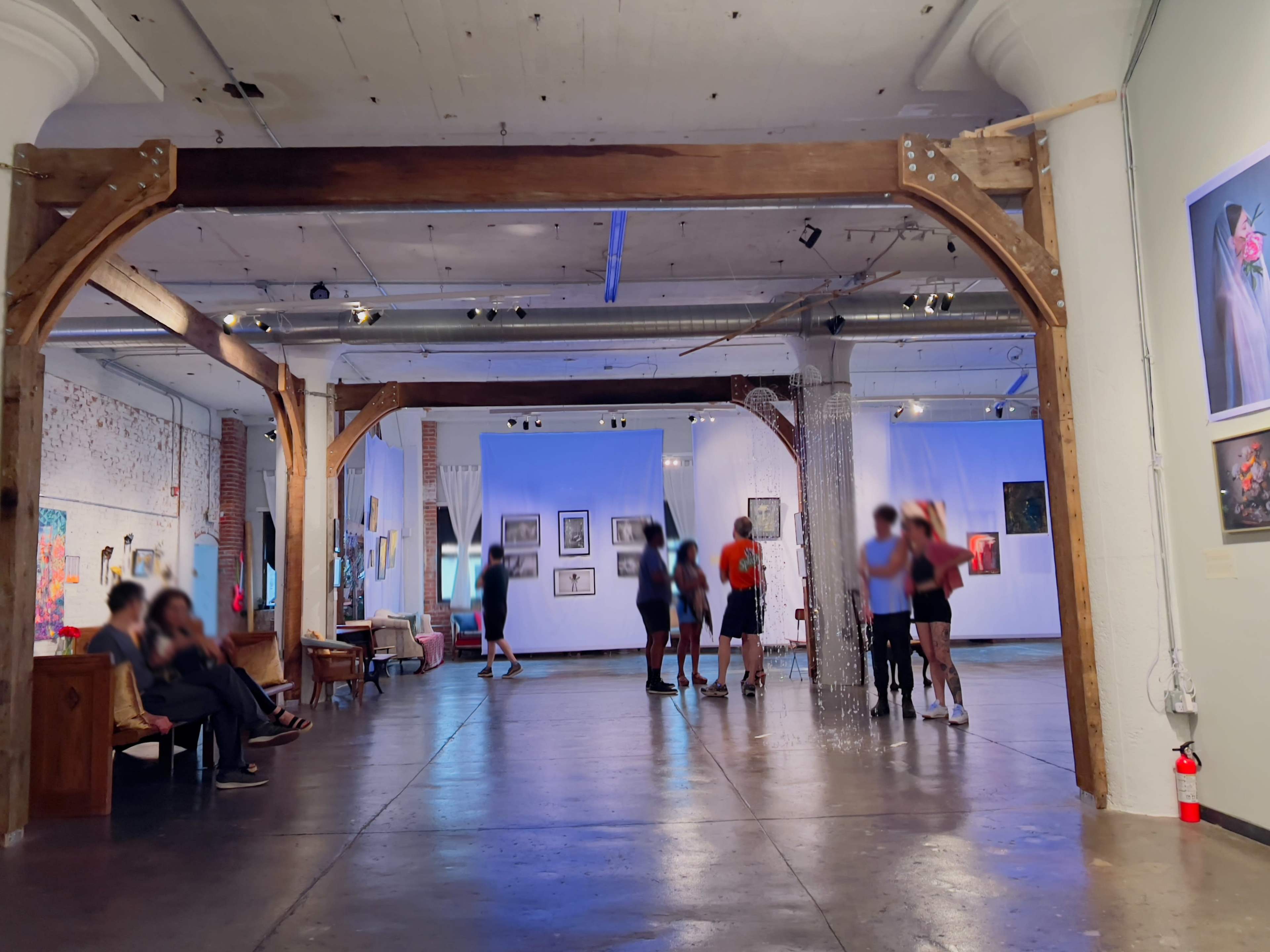 An art gallery with wooden beams, people engaging in activities, and various artworks displayed on the walls.