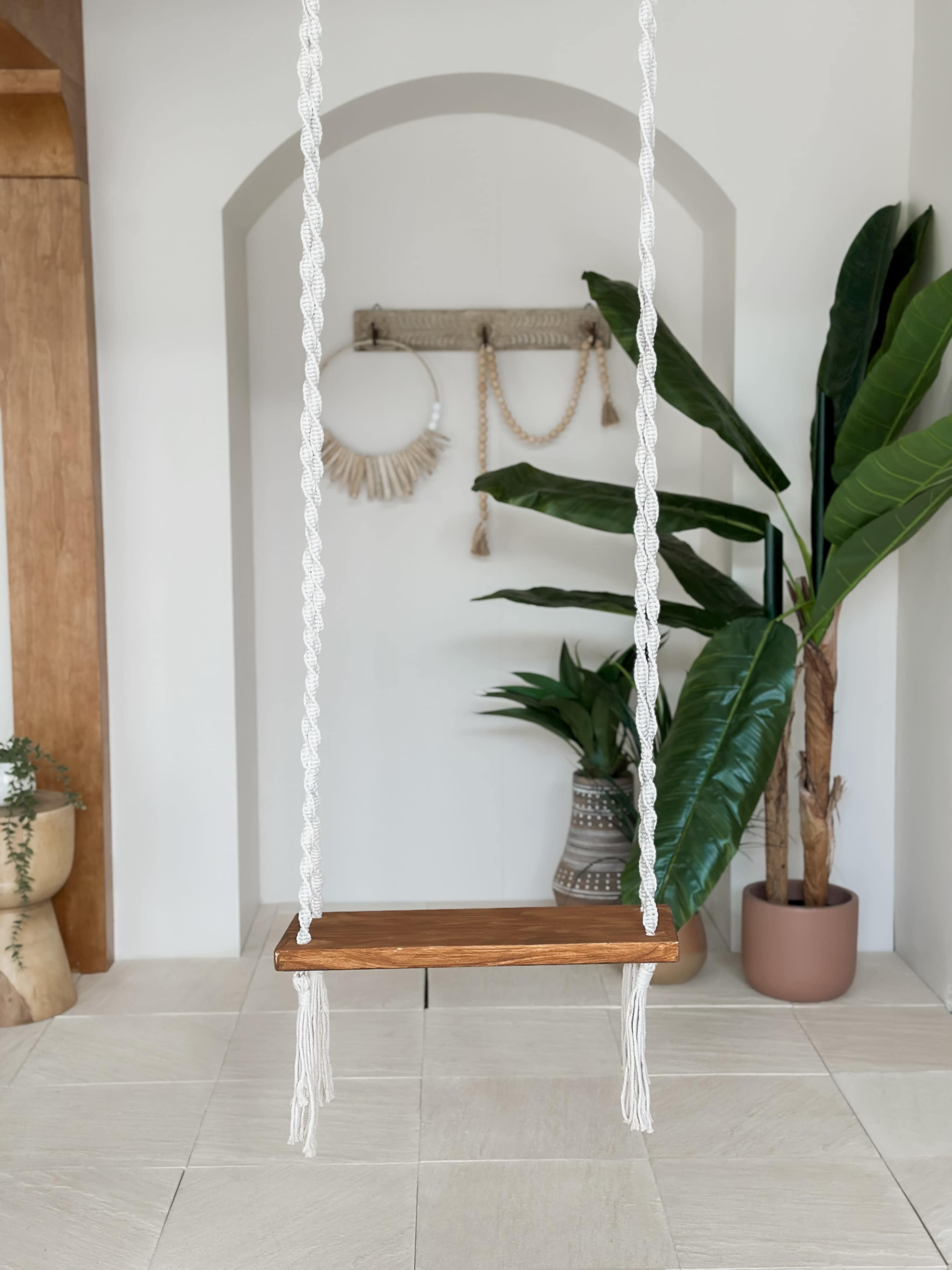 A wooden swing hangs from braided ropes in an indoor space adorned with plants and decorative wall hangings.