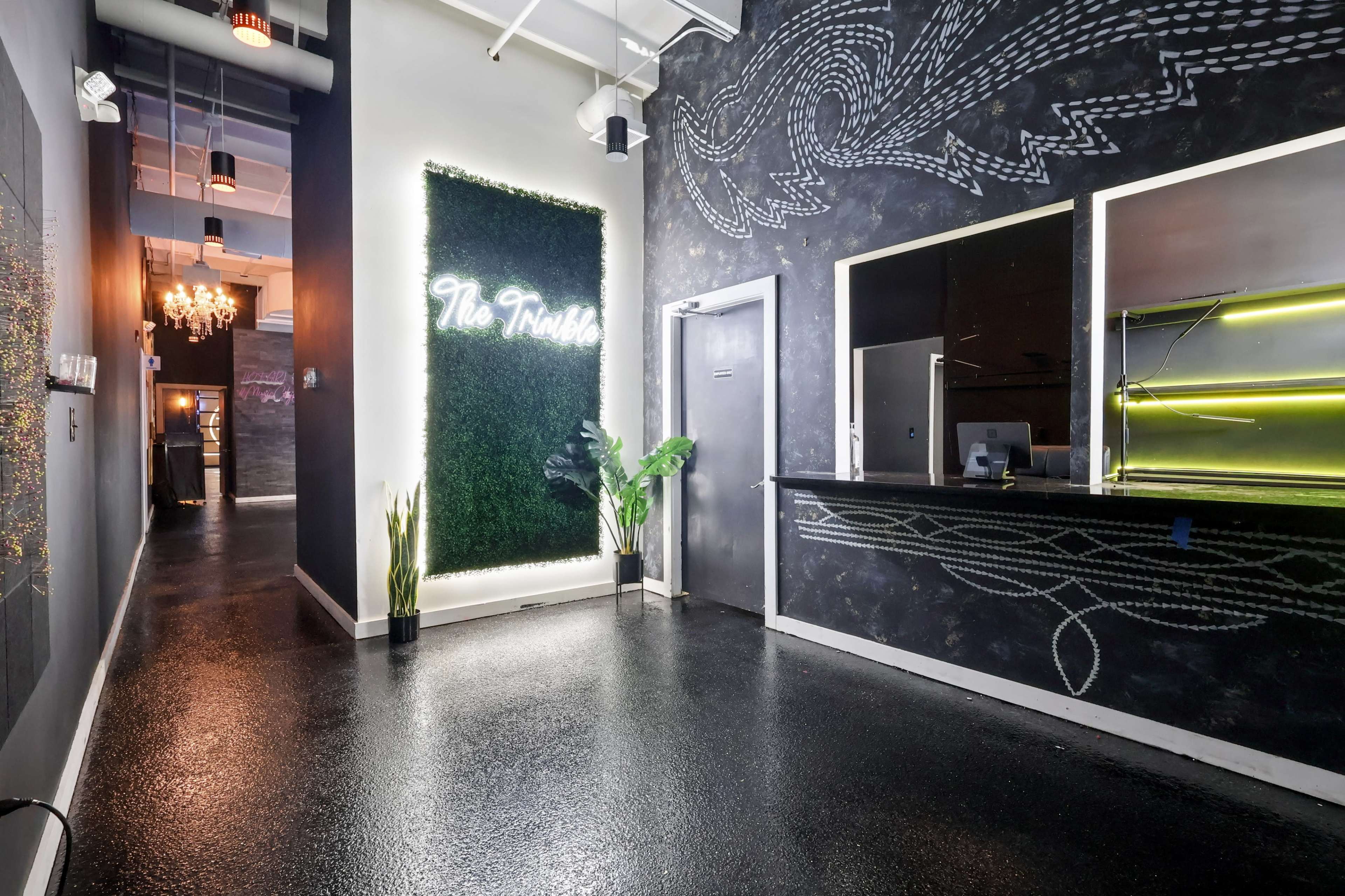 The image shows a modern, stylish reception area with a dark wall featuring a neon sign that reads "The Trotto," alongside a sleek counter and decorative plant.