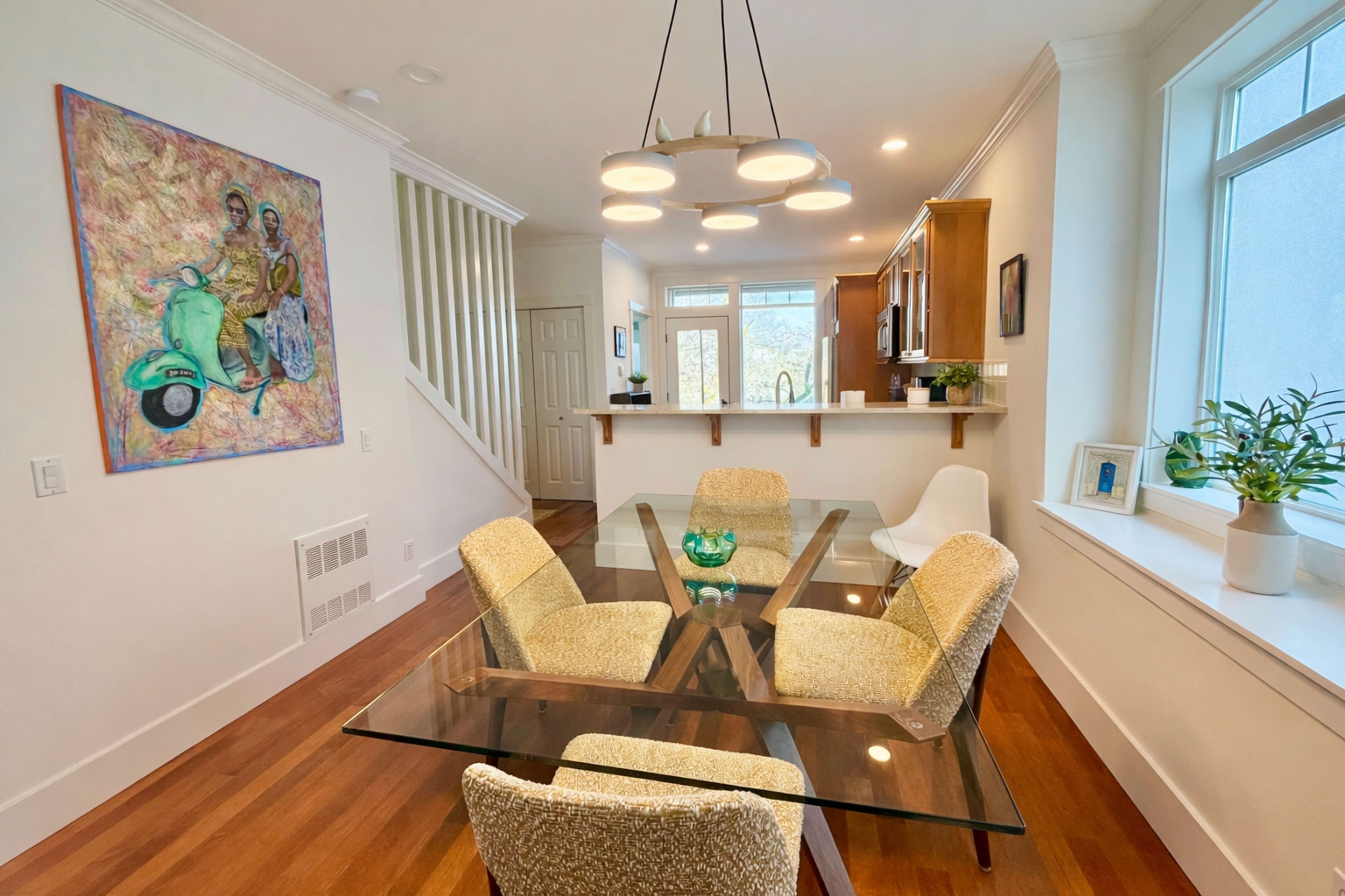 The image shows a modern dining area featuring a glass table surrounded by four chairs, with an open kitchen space in the background and a colorful painting on the wall.