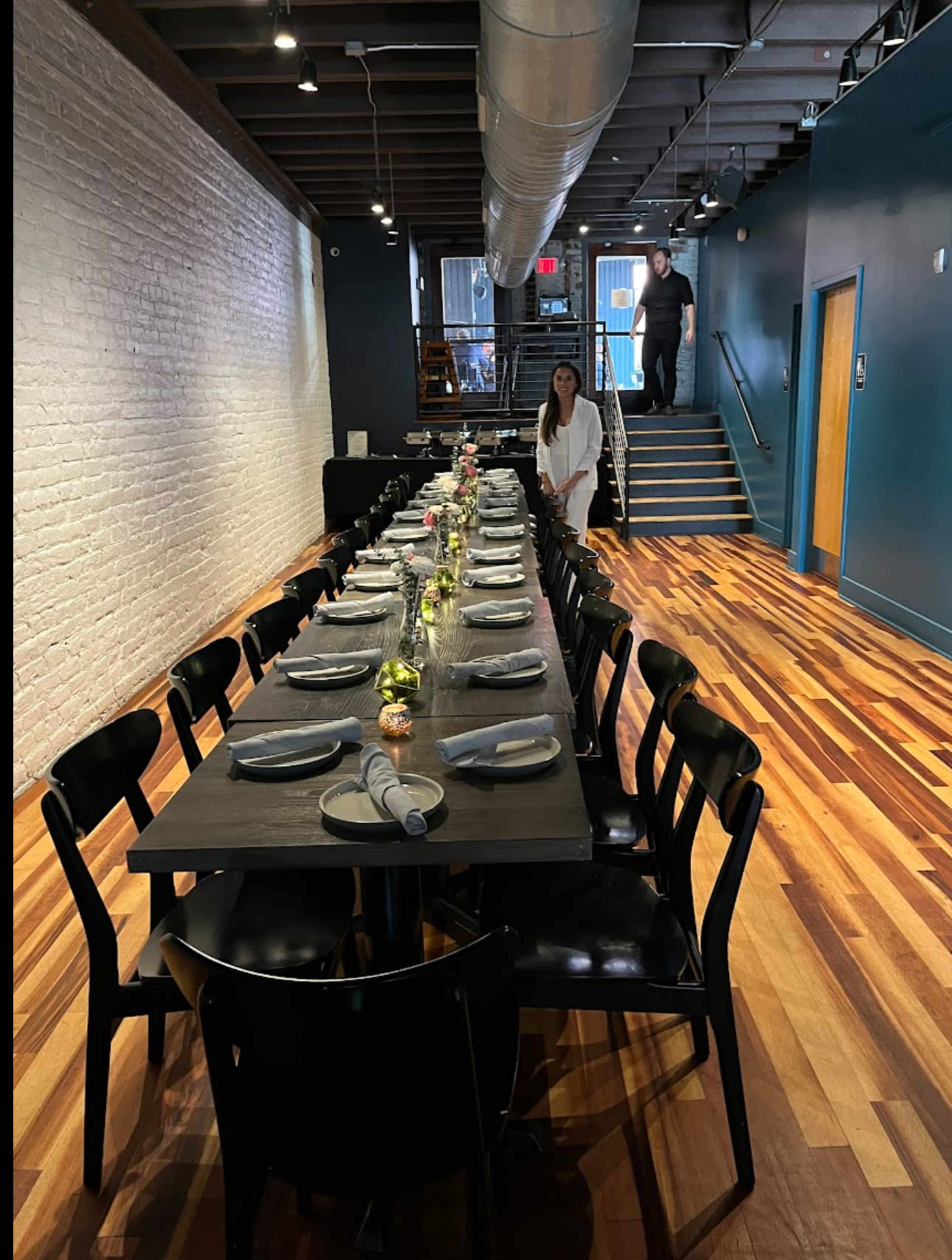 A long dining table is set with plates and utensils, while a woman stands near the table, and a person walks up a staircase in the background.