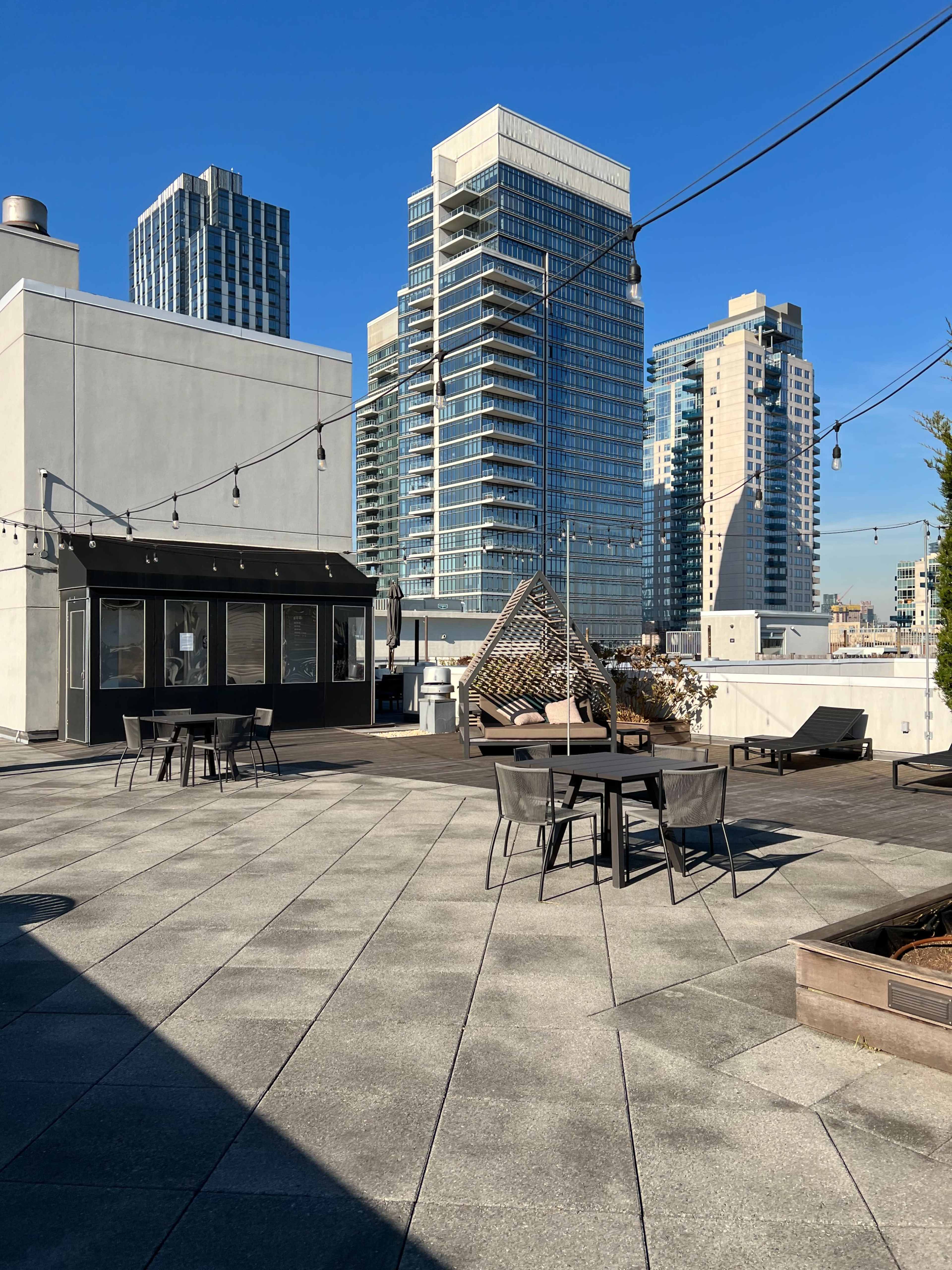 A rooftop patio with seating areas, string lights, and tall buildings in the background under a clear blue sky.