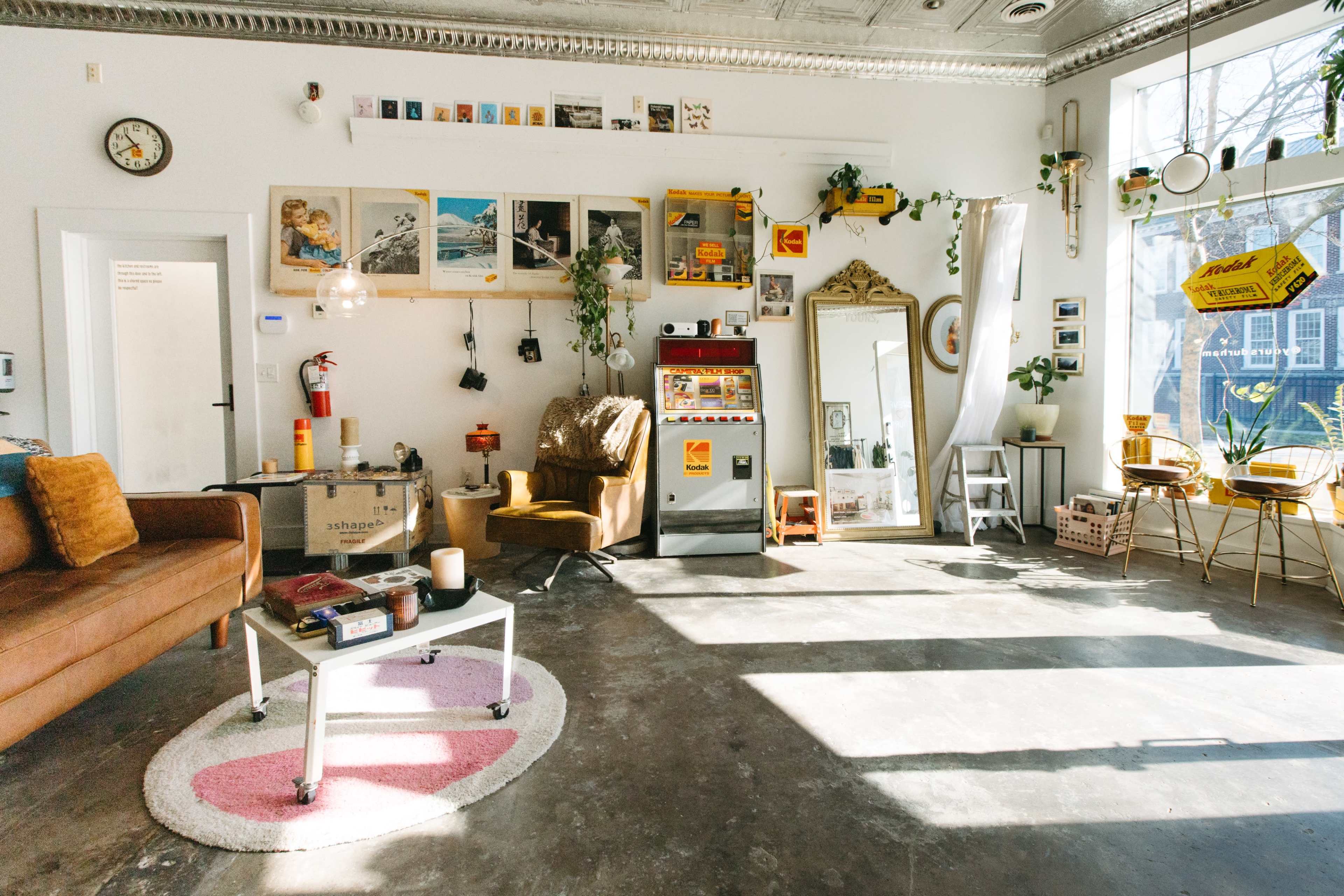 The image shows a cozy indoor space with a brown sofa, a vintage armchair, art on the walls, and various plants arranged throughout.