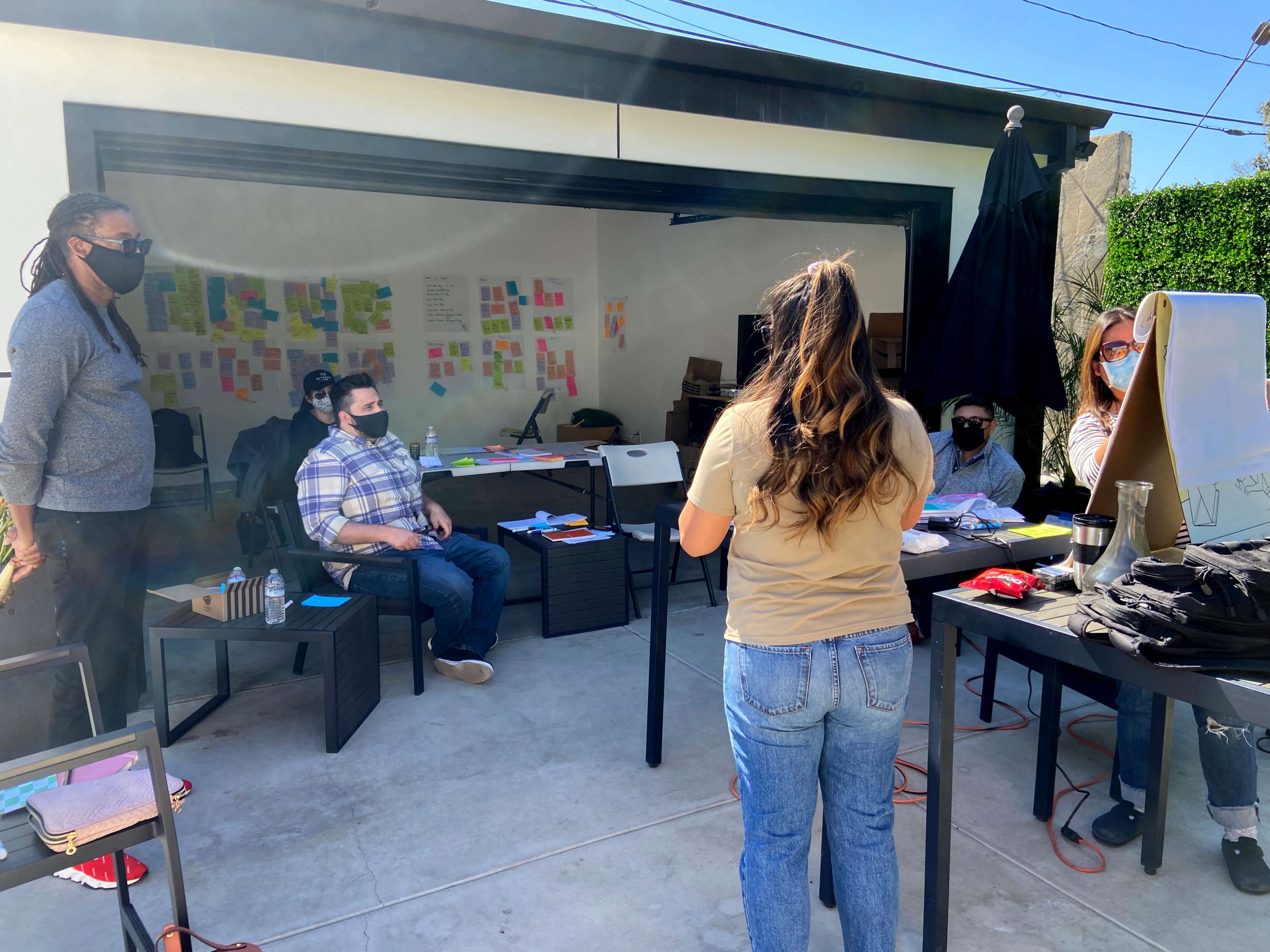 A group of people wearing masks is engaged in a meeting outdoors, with sticky notes displayed on a wall in the background.