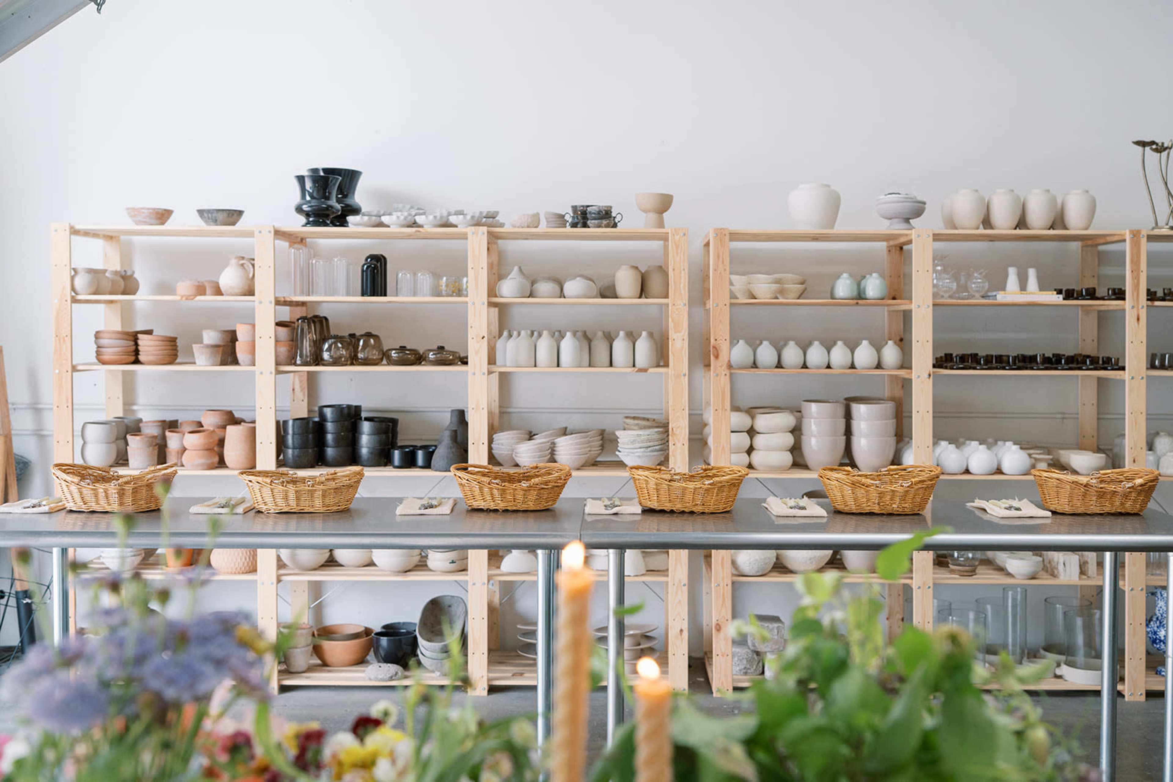 The image shows a spacious pottery studio with wooden shelves filled with an assortment of ceramic items and several wicker baskets placed on tables in front.