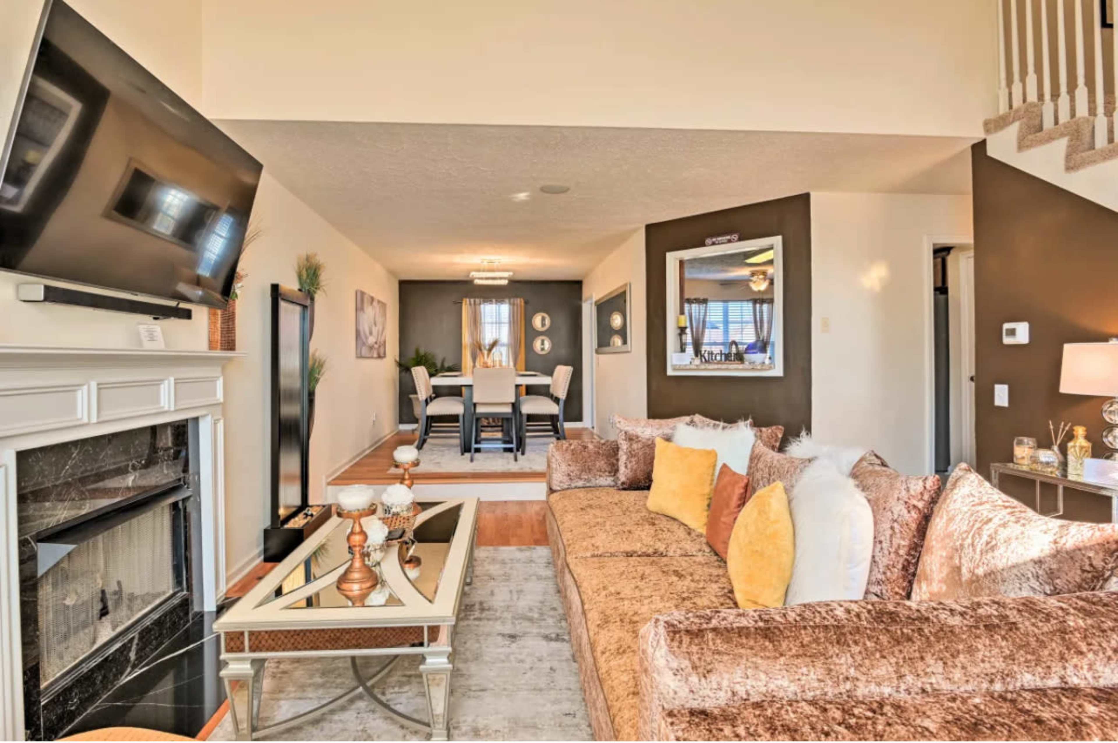 A modern living room features a plush sofa, a television mounted above a fireplace, and a view of a dining area through an archway.