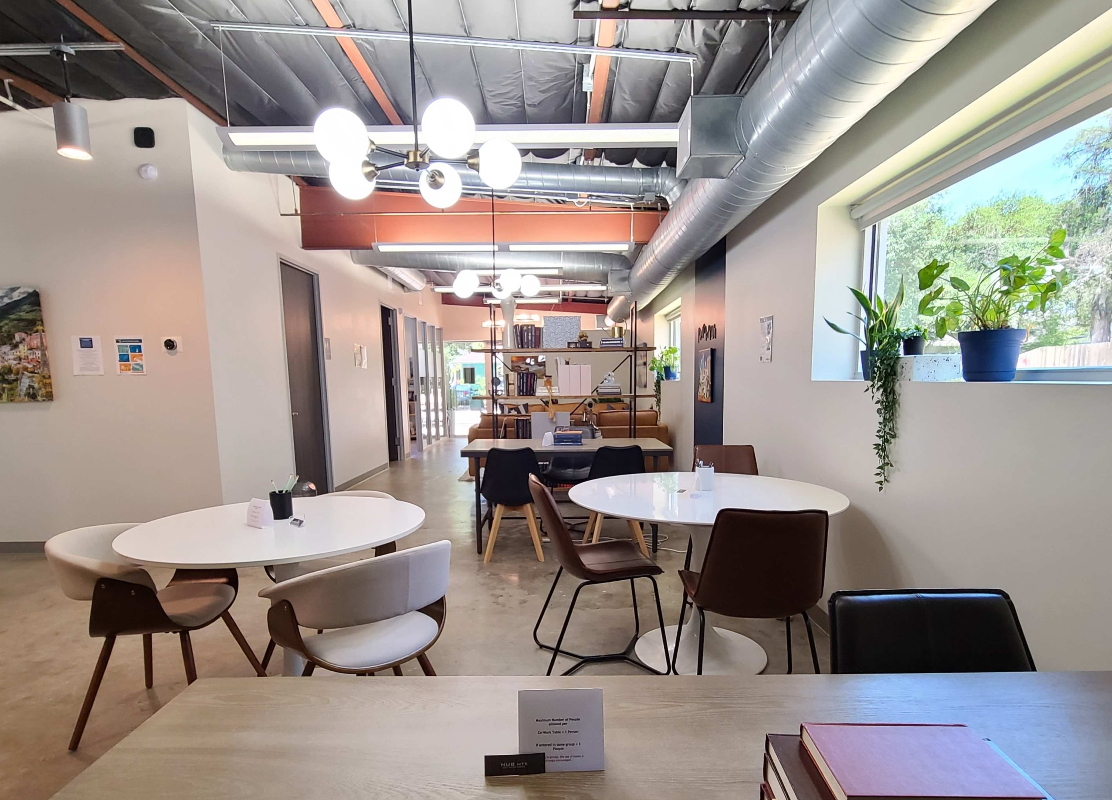 The image shows a modern co-working space with several white and wooden tables surrounded by chairs, and plants placed in the windows.
