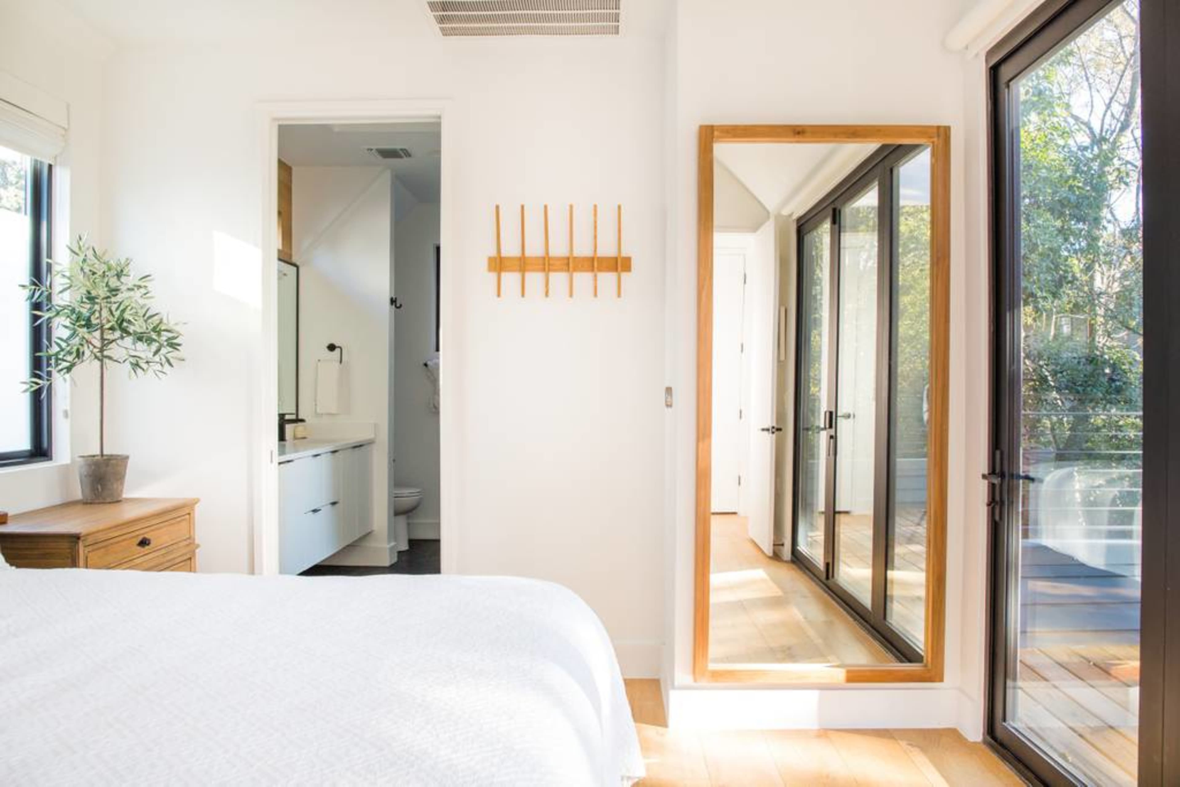 A bright bedroom features a bed, a large mirror, and a doorway leading to a bathroom.