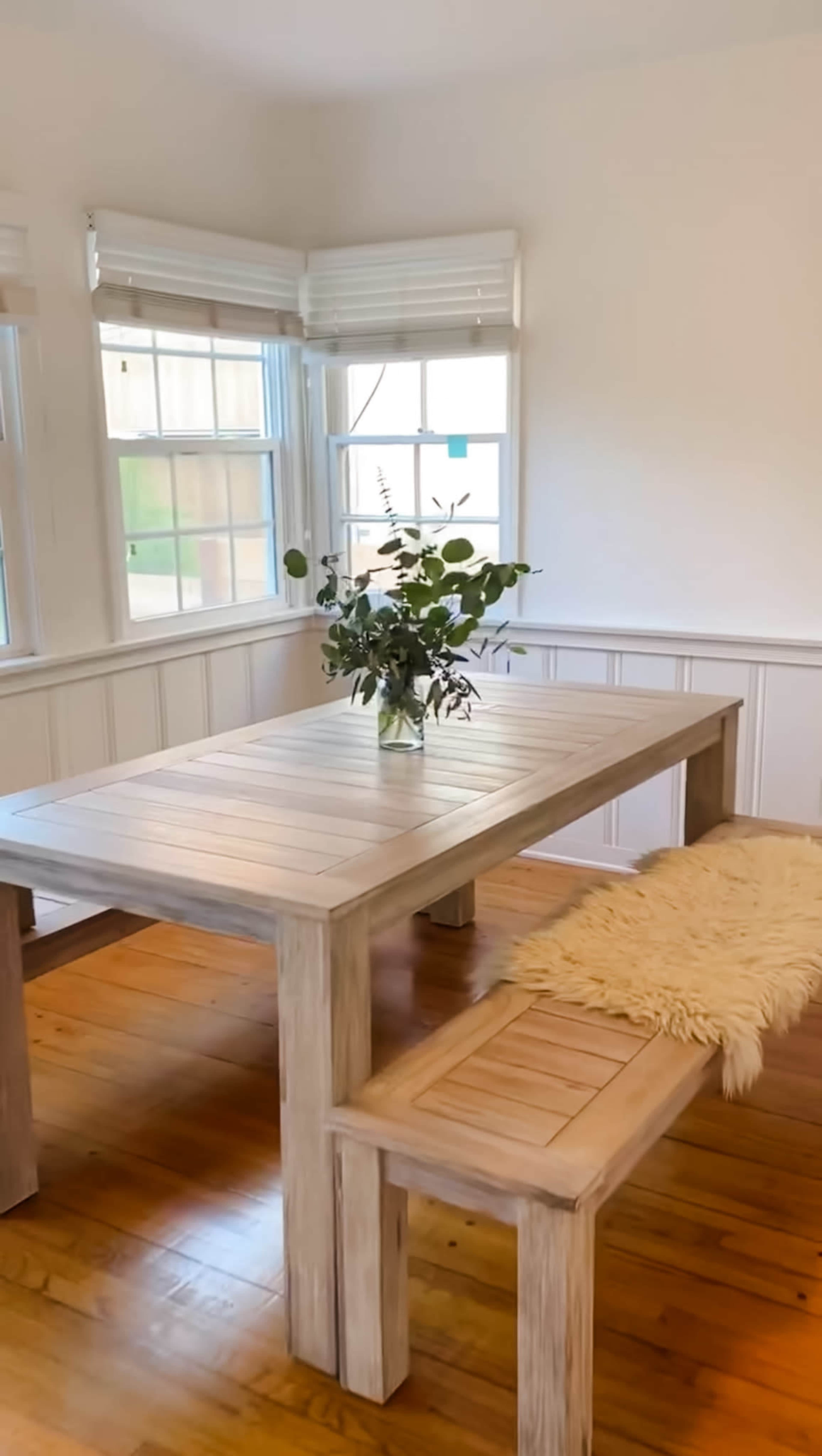 A wooden dining table with a bench is positioned in a well-lit room, adorned with a vase of greenery.