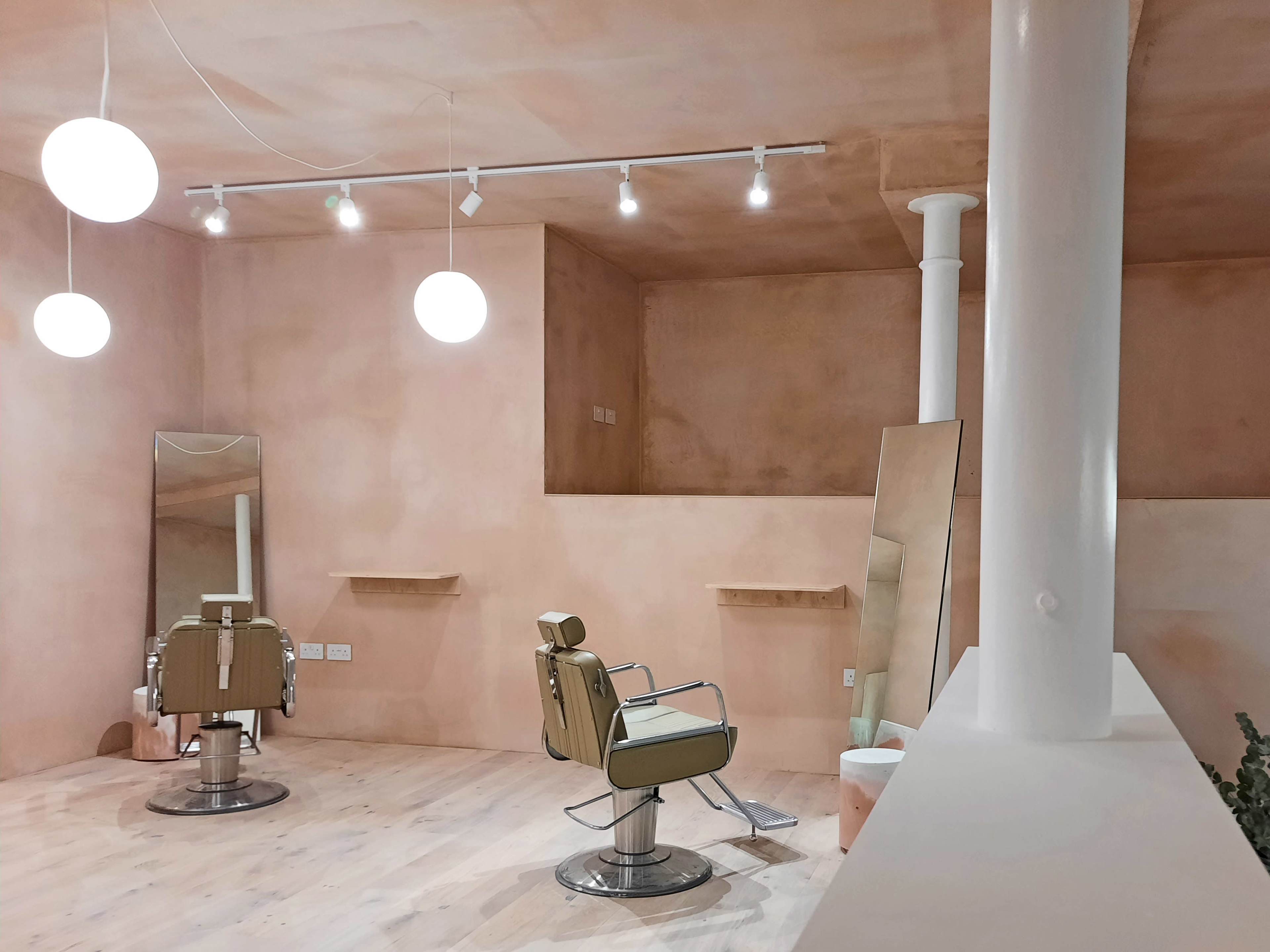 The image shows a minimalist hair salon with two styling chairs and large mirrors against a softly colored, smooth wall.