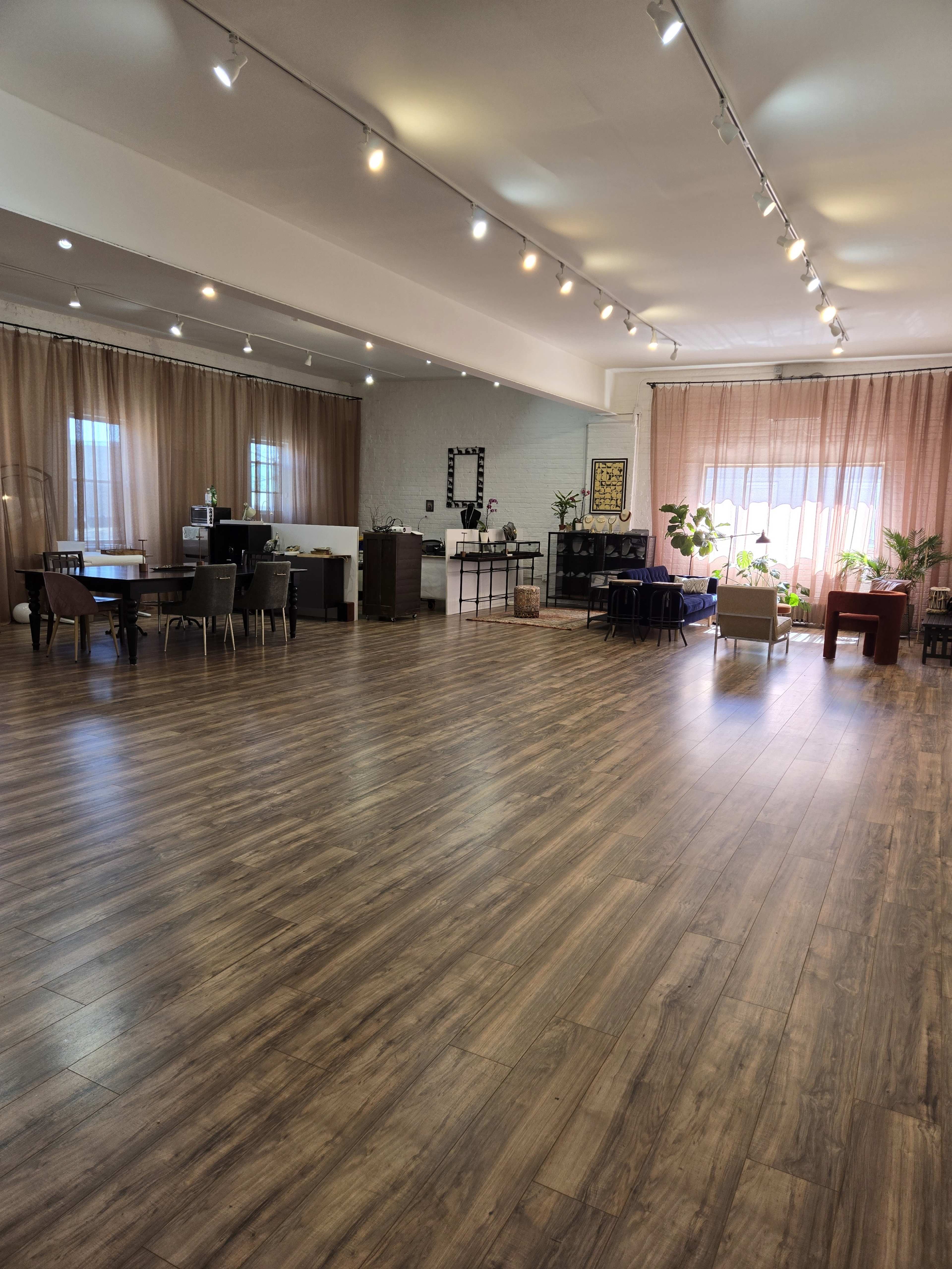 A spacious interior with wood-like flooring, featuring various seating areas, plants, and large windows with sheer curtains.