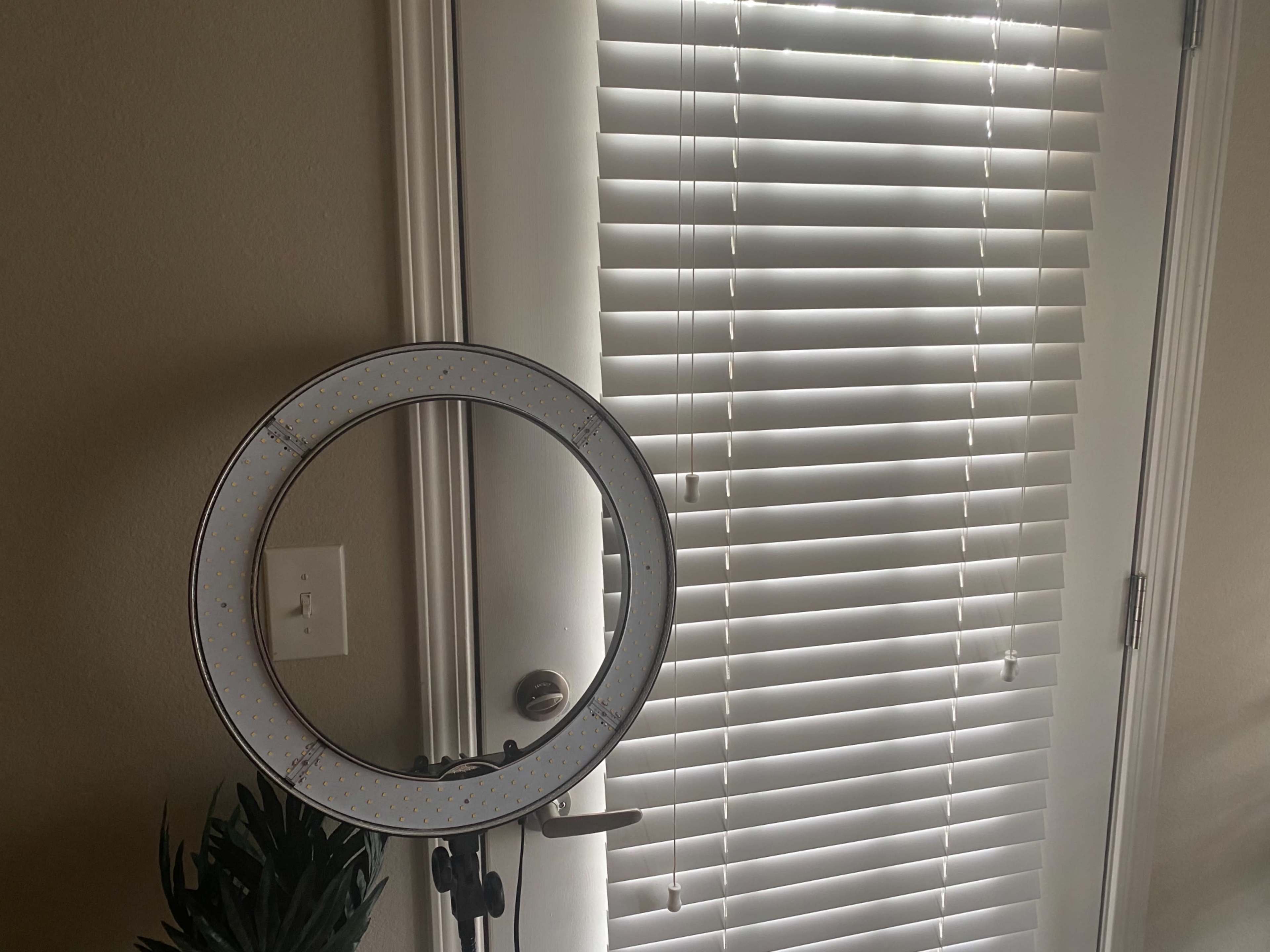 A ring light stands in front of a partially open door with blinds.