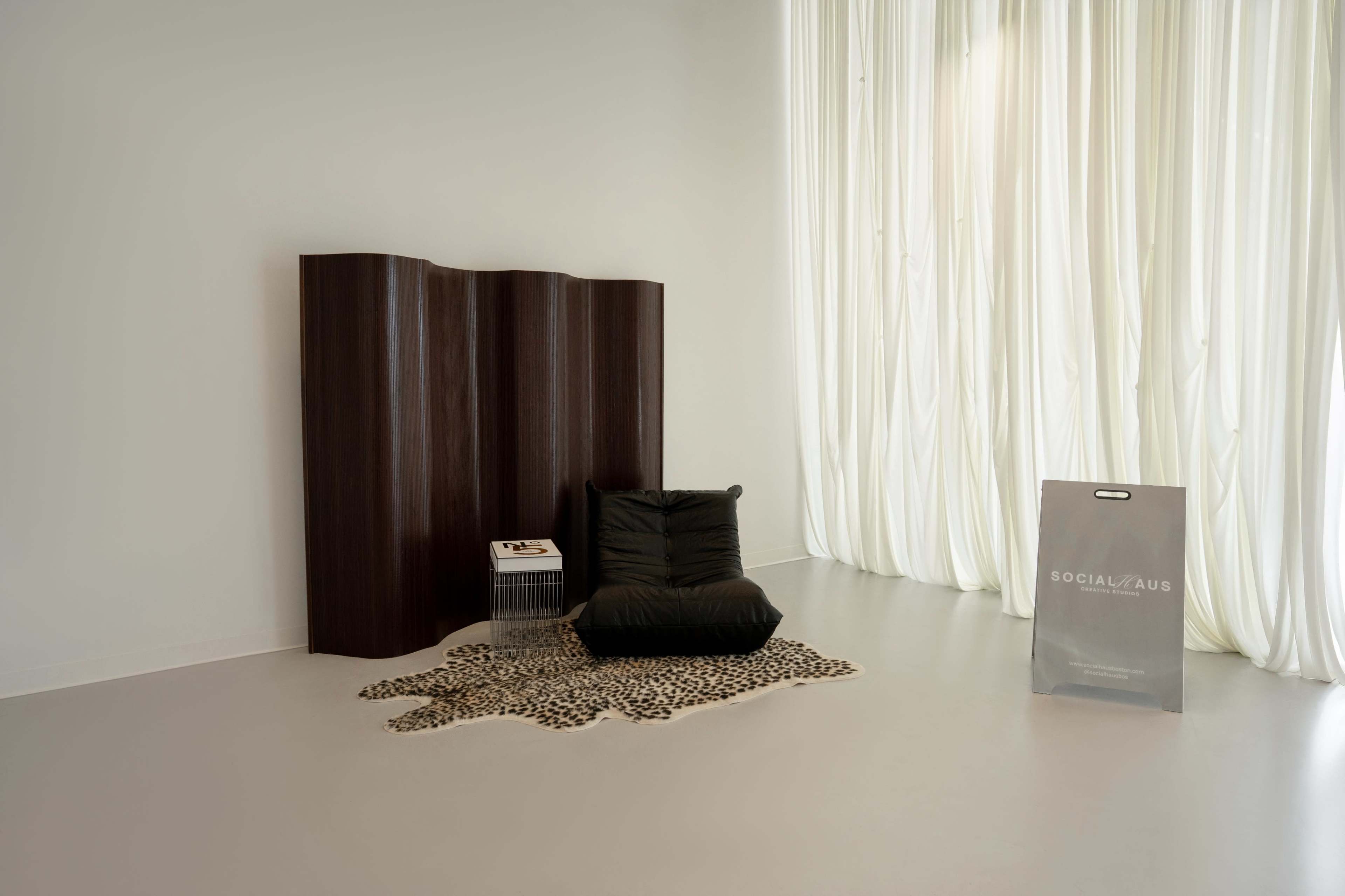 The image shows a minimalist room featuring a wooden partition, a black beanbag chair on a leopard print rug, and a large bag labeled "SOCIAL/AUS" near sheer white curtains.