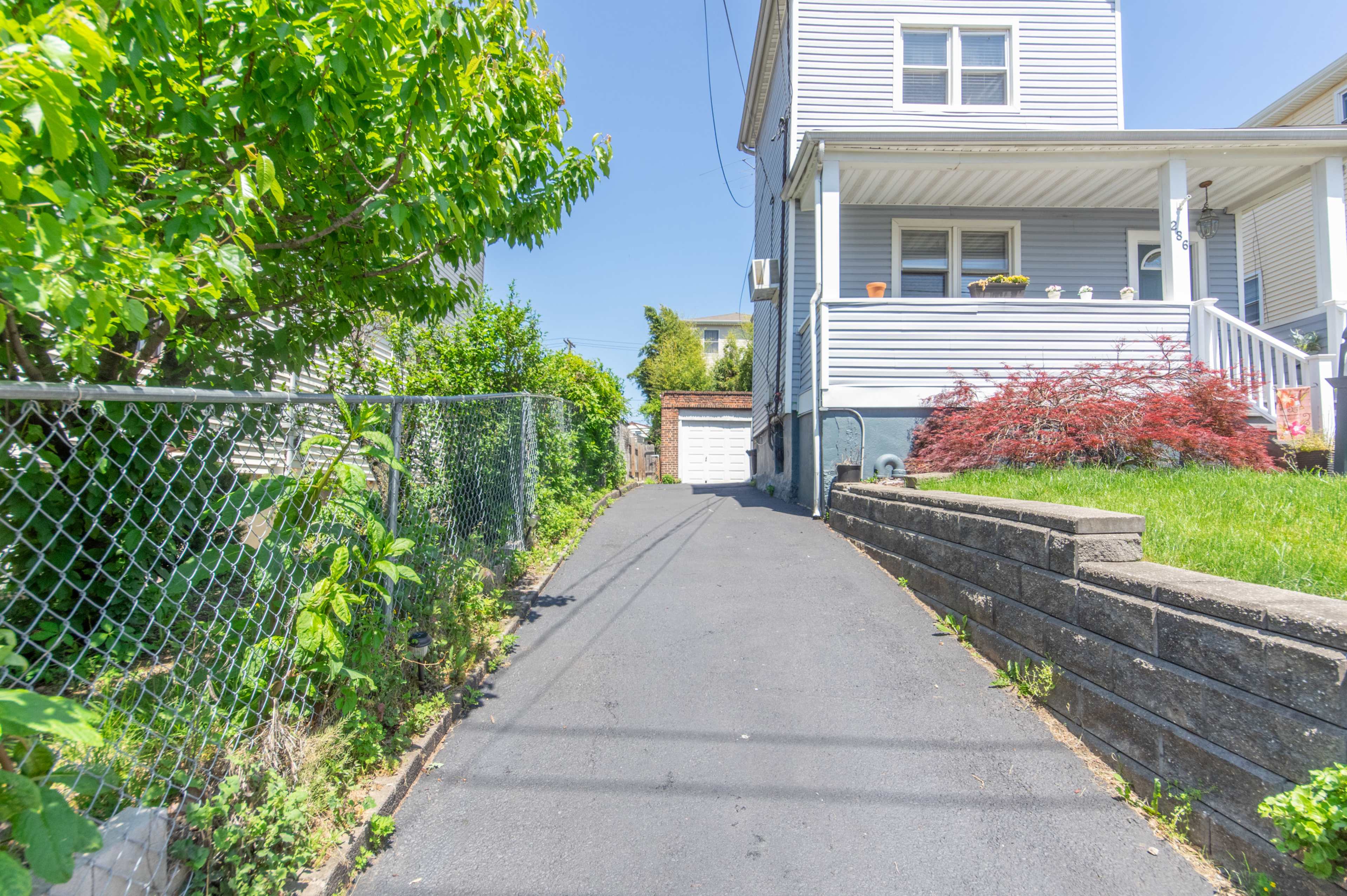 A narrow asphalt driveway lined with greenery leads to a white garage at the back of a residential property.