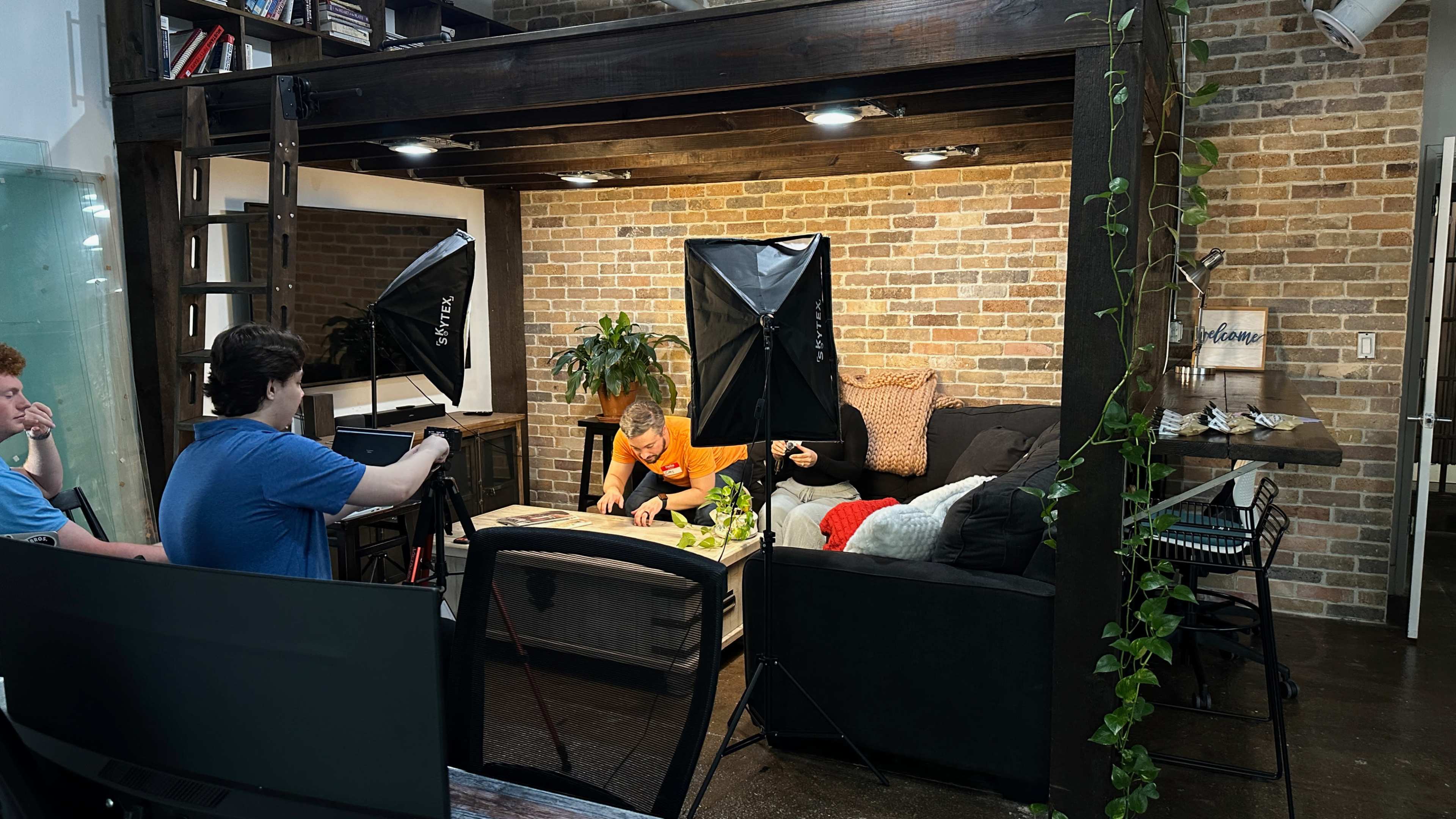 A small production set features a man and a woman working at a table in a cozy living room space, with lighting equipment arranged around them.