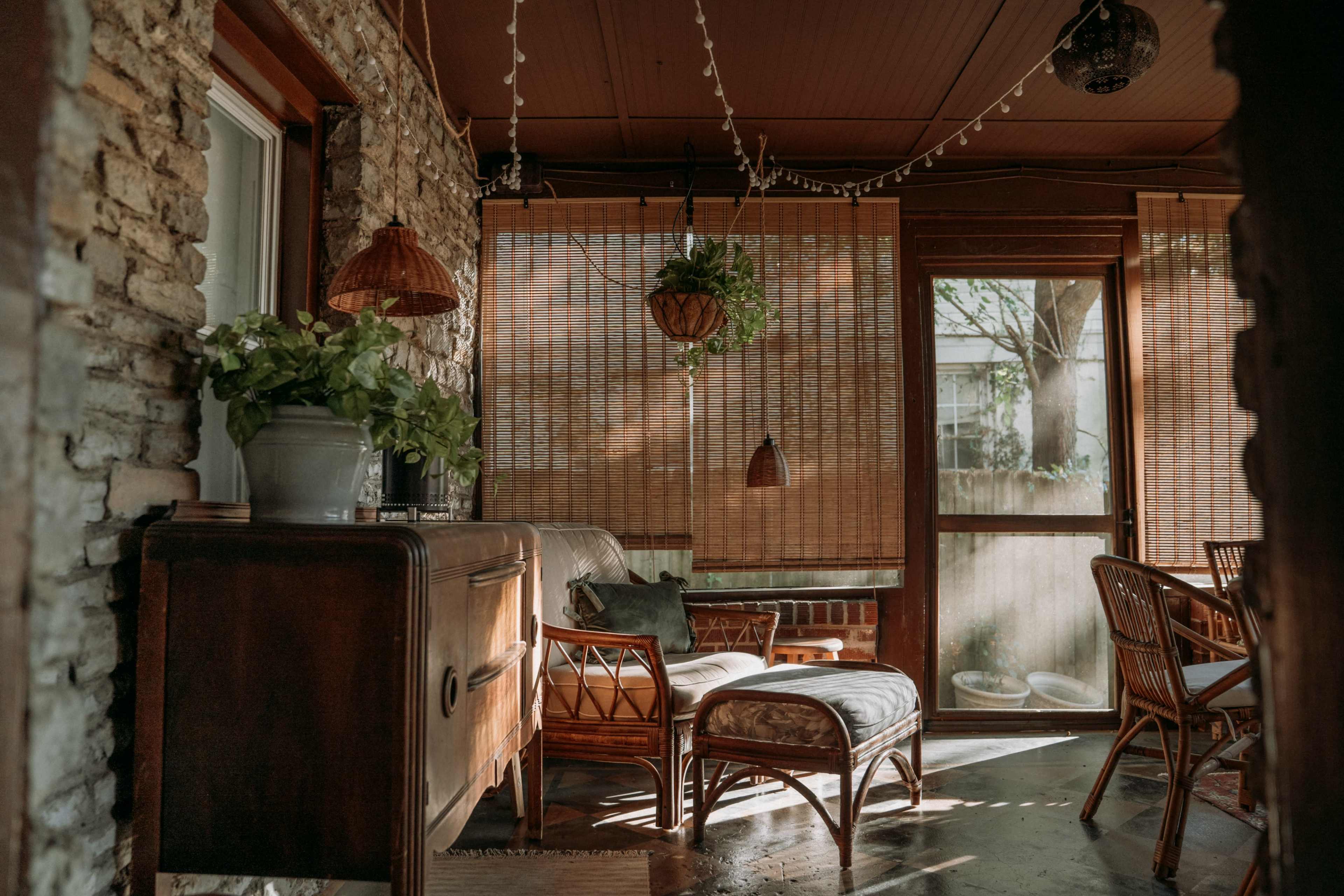 The image features a cozy sunroom with wooden furniture, potted plants, and bamboo shades allowing natural light to filter in.