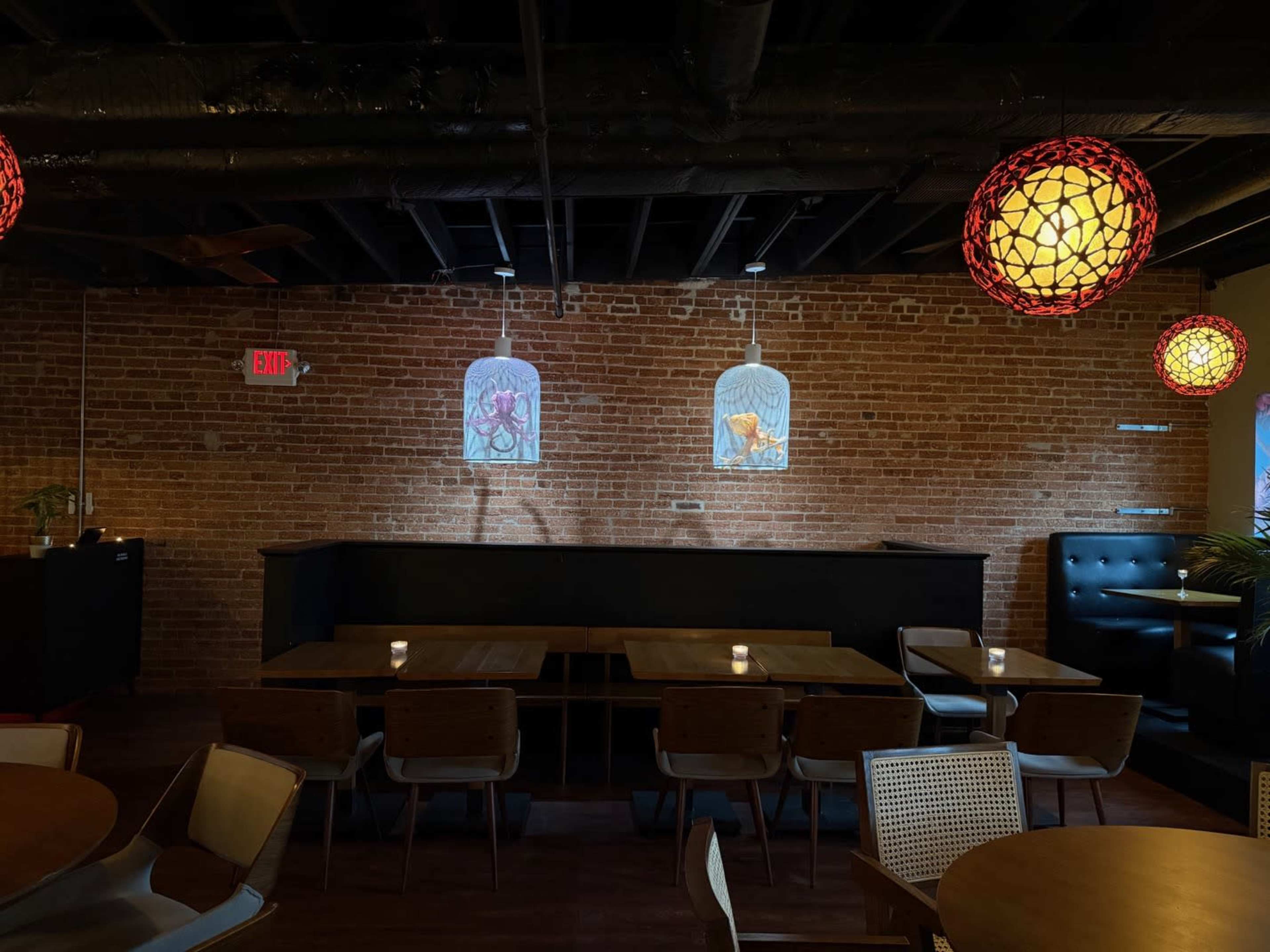 A dimly lit restaurant features wooden tables and modern chairs, with exposed brick walls and colorful pendant lights.