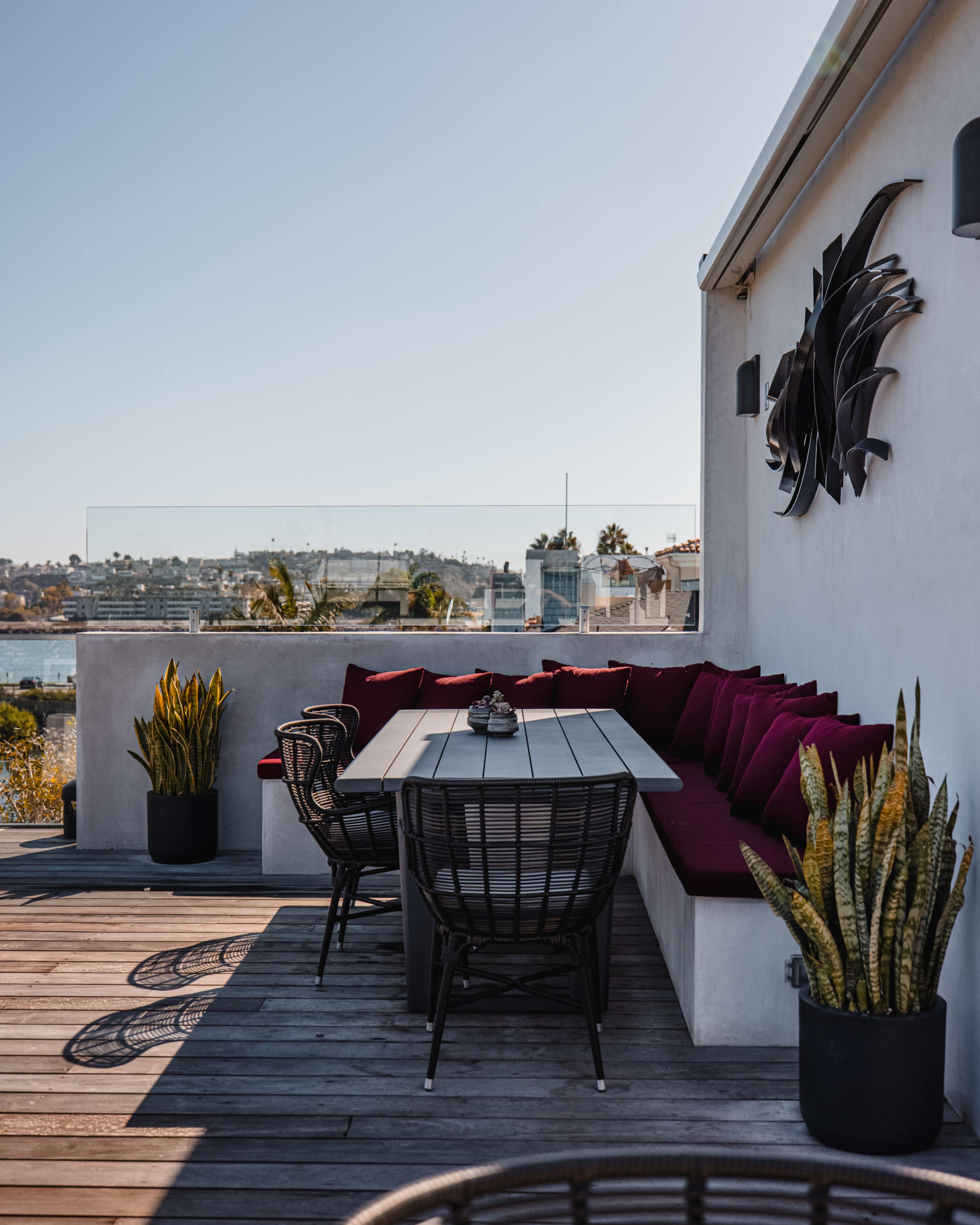 A modern outdoor dining area features a long table surrounded by black wicker chairs and a cushioned bench against a scenic backdrop of water and buildings.