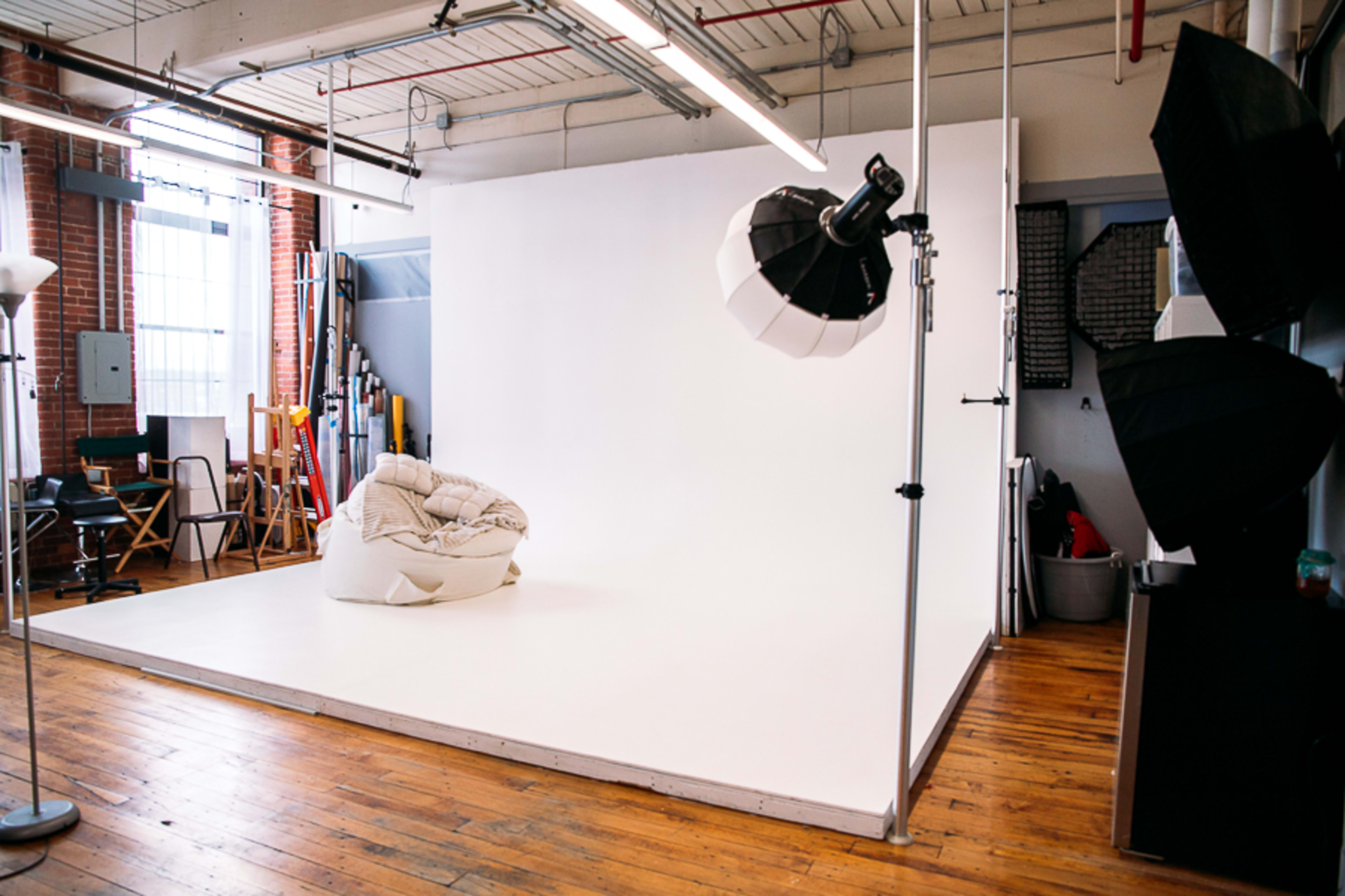 The image shows a photography studio with a white backdrop, a bean bag chair in the center, and various lighting equipment set up around the room.