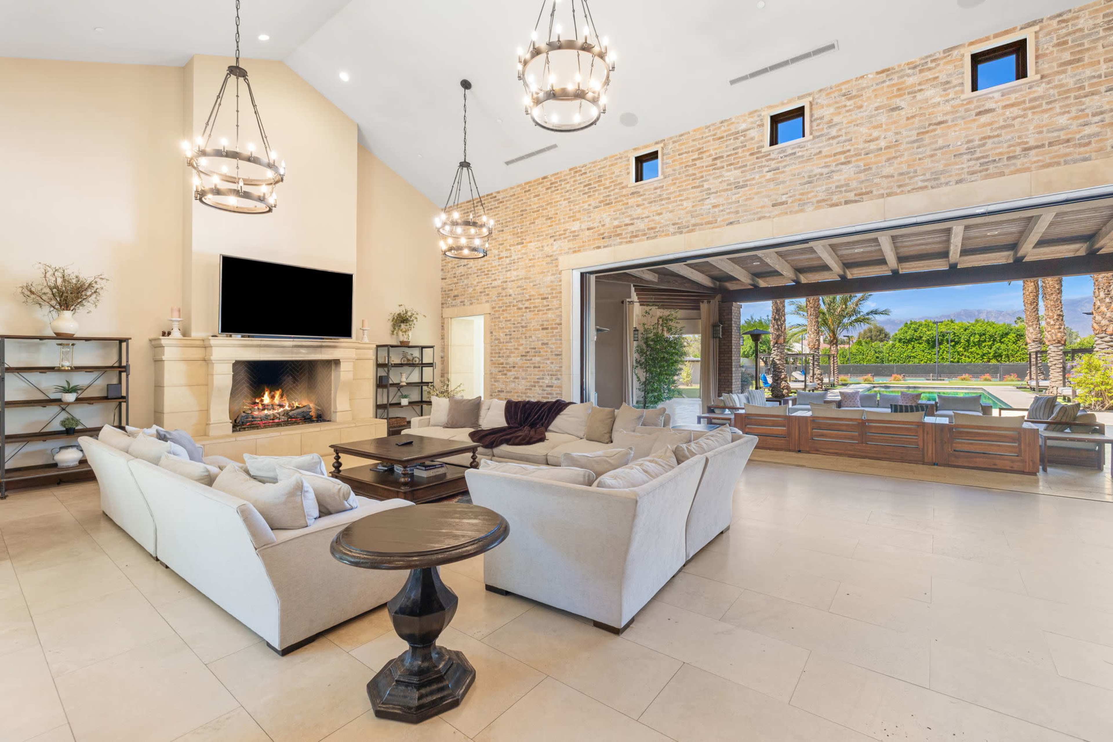 A spacious living room features a large sofa, a coffee table, and a fireplace, with large sliding doors opening to an outdoor area with a pool and palm trees.