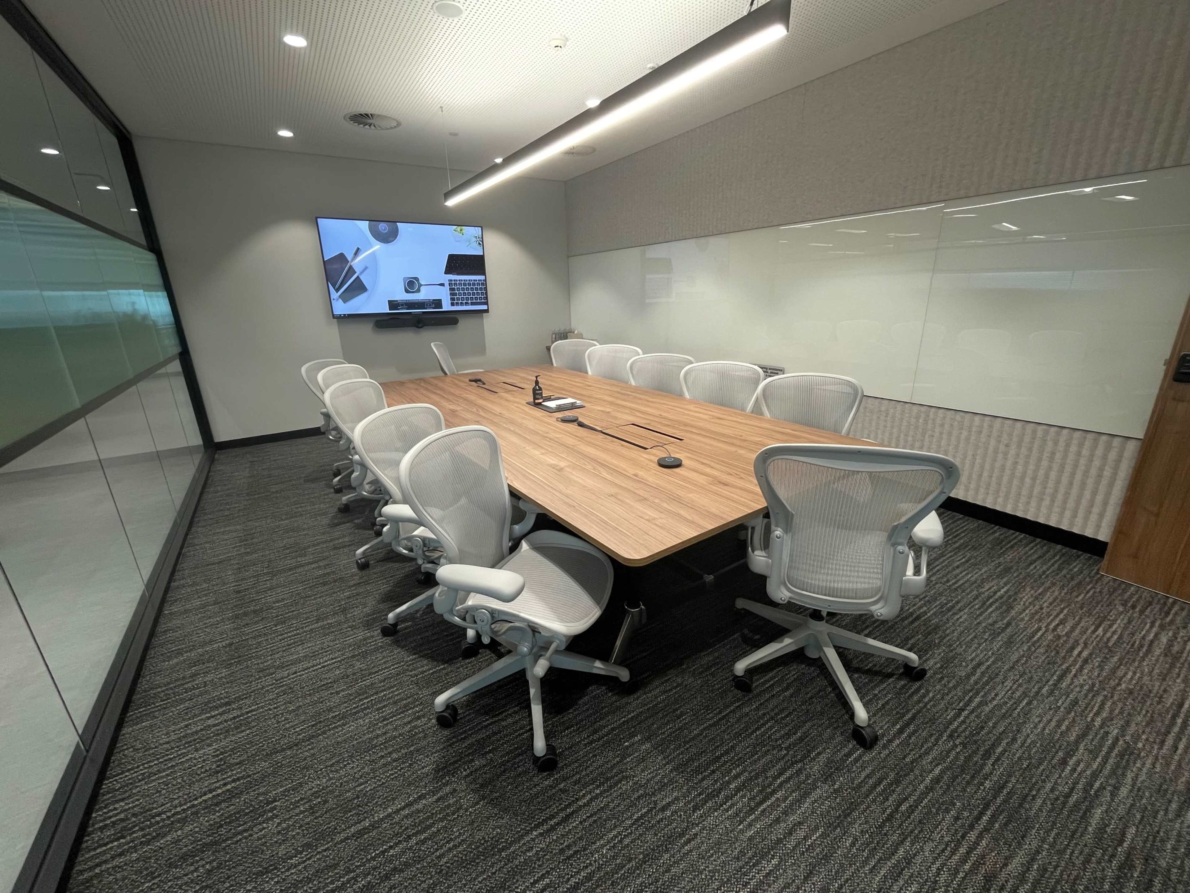 A modern conference room features a large wooden table surrounded by several ergonomic chairs and a wall-mounted screen.