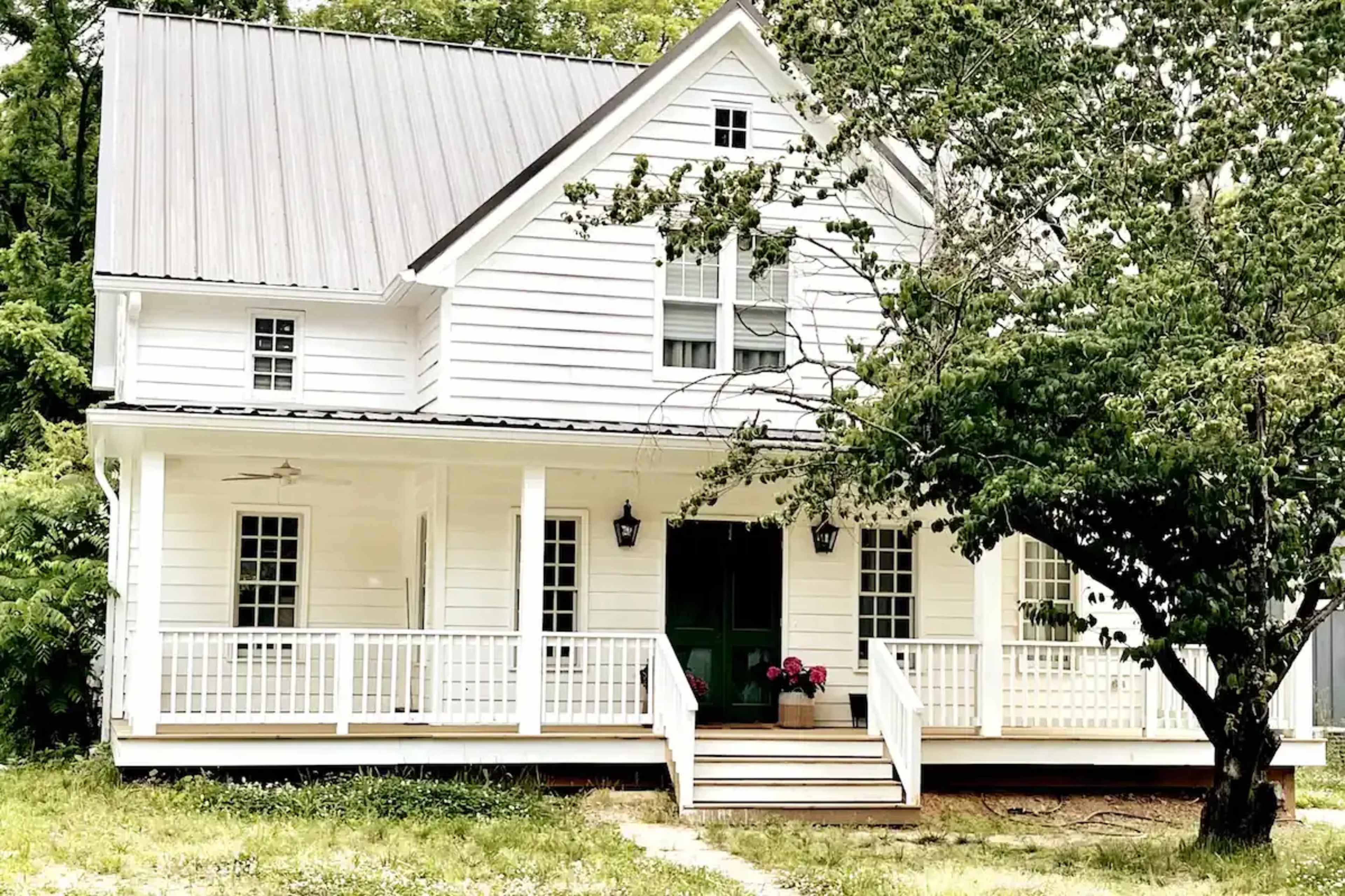 A white, two-story house with a metal roof features a front porch and green door, surrounded by trees and a flower planter.