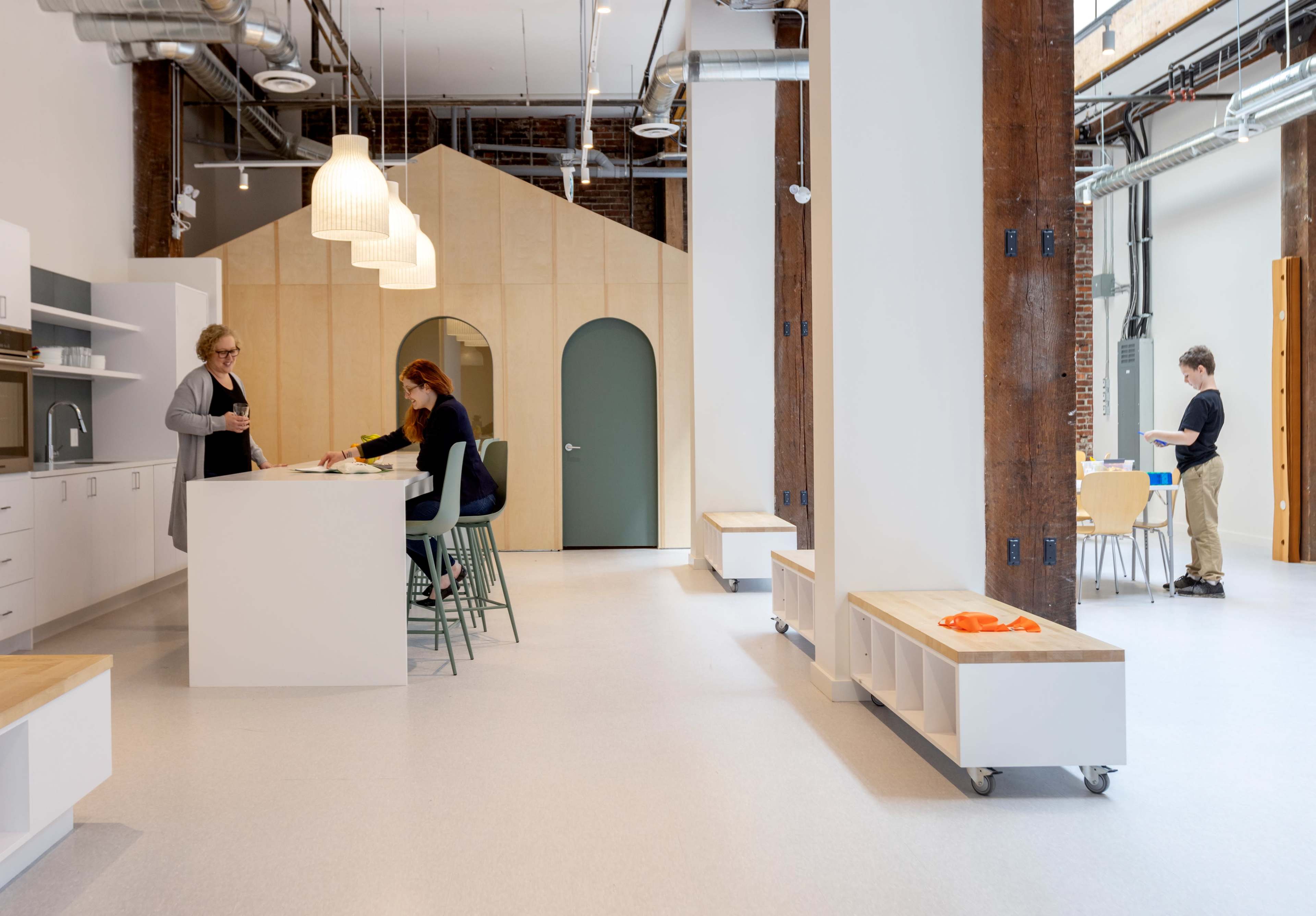 A modern, open-concept workspace with two women preparing food at a central island and a man setting up tables in the background.